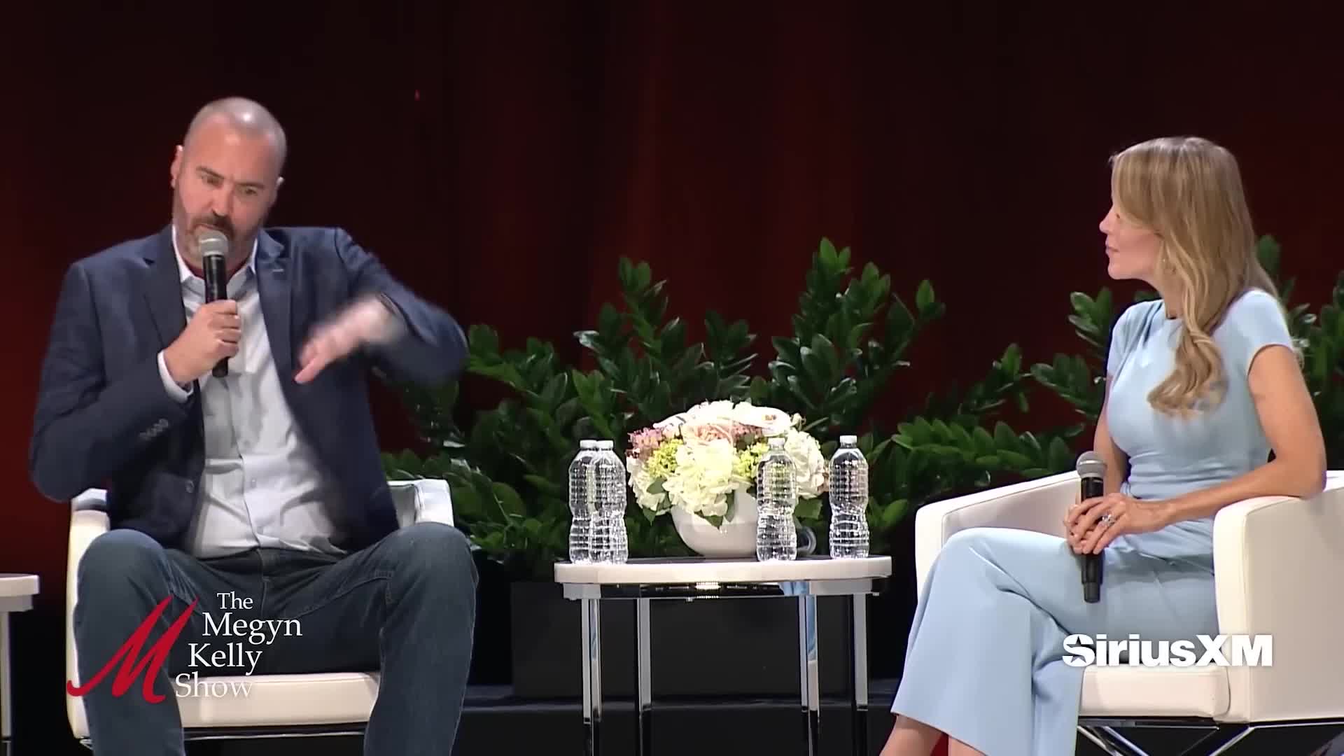 The man, holding a microphone, gestures expansively while speaking. Across from him, a woman in a powder blue outfit listens, also holding a microphone.
