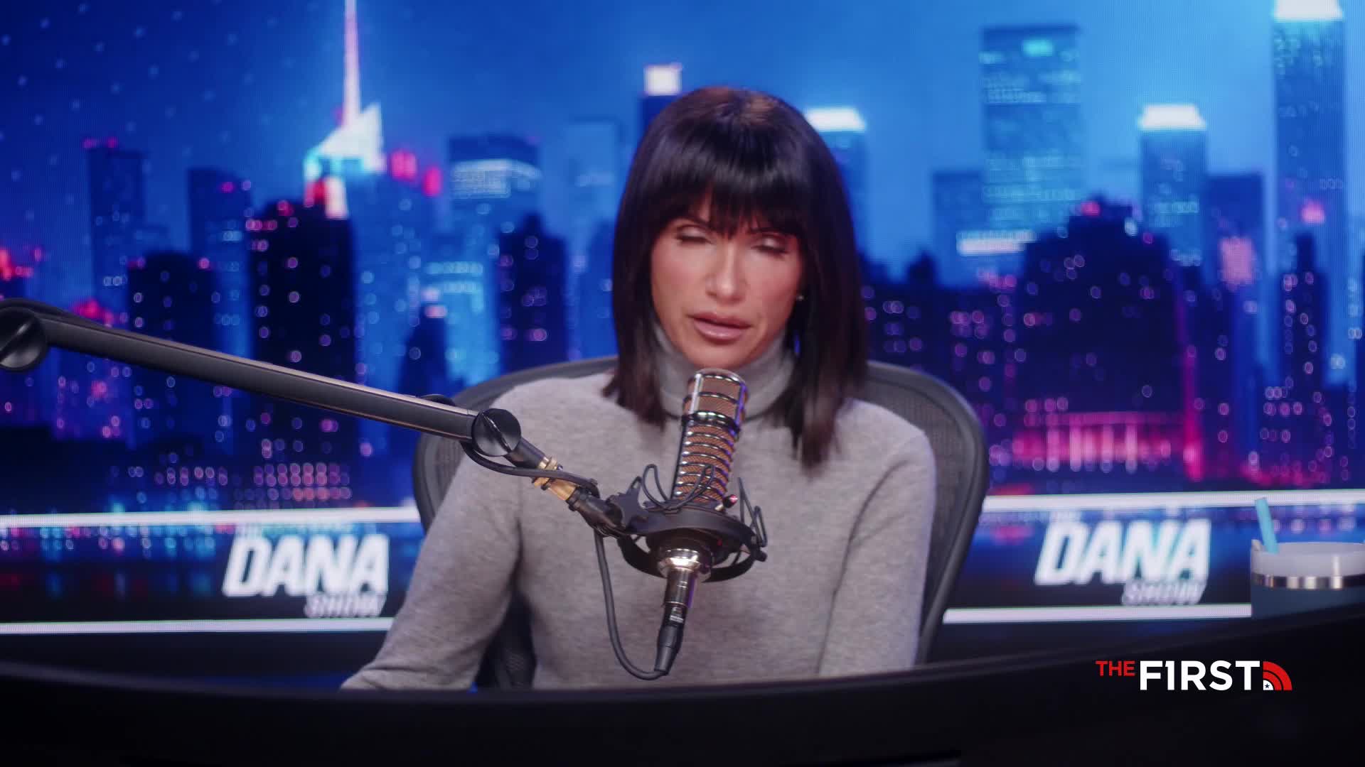 The host of "The Dana Show" on The First TV is speaking into a microphone. Behind her, a backdrop depicts a nighttime cityscape in the United States.
The host of "The Dana Show" on The First TV is speaking into a microphone. Behind her, a backdrop depicts a nighttime cityscape in the United States.