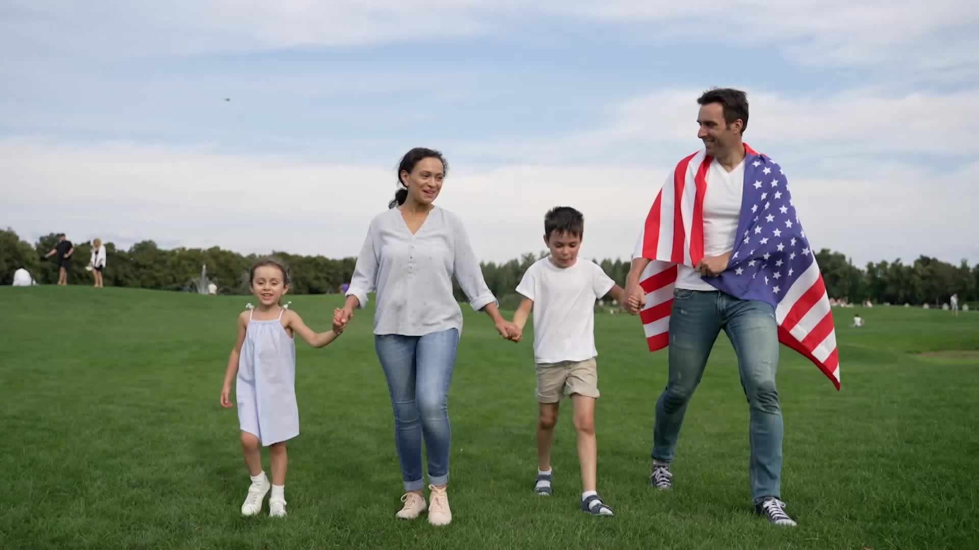 A family of four walks across a grassy field, holding hands. The father has an American flag draped over his shoulders as they stroll.
A family of four walks across a grassy field, holding hands. The father has an American flag draped over his shoulders as they stroll.