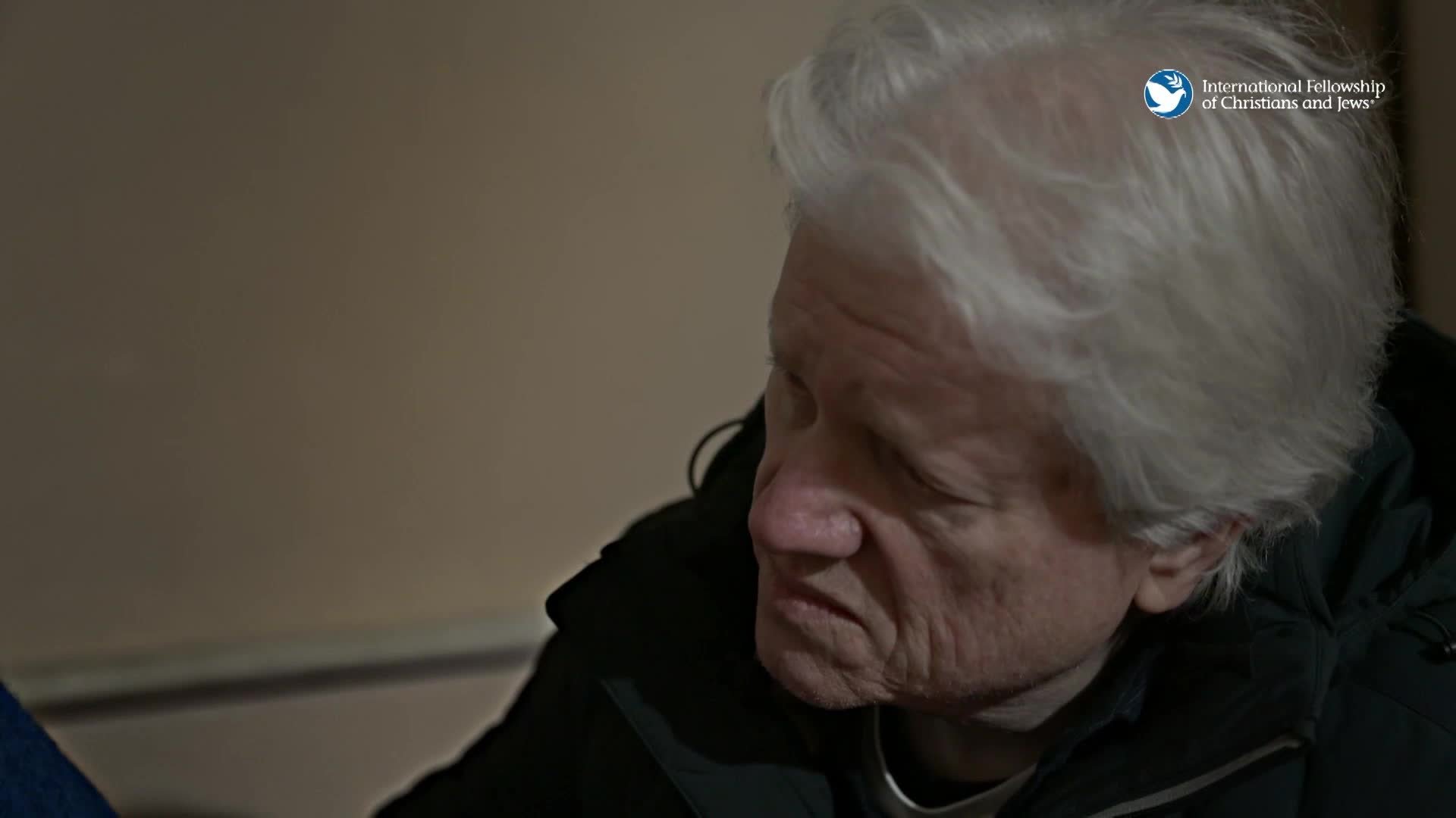 A man with white hair sits, his head bowed slightly. He wears a dark jacket, and the light catches the lines on his face.