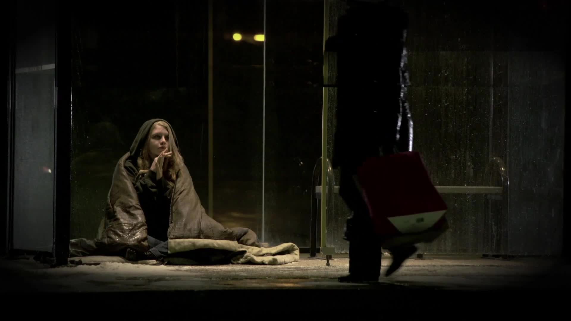 A young woman, wrapped in a blanket, sits on the ground at a bus stop. A figure walks past, carrying a red bag.