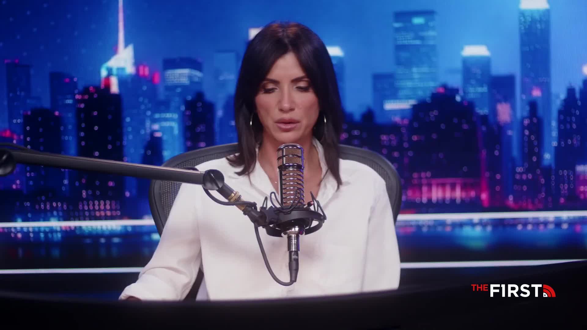 A woman in a white shirt speaks into a microphone, her dark hair framing her face. Behind her, a cityscape glows with neon lights, hinting at a broadcast from The First TV.