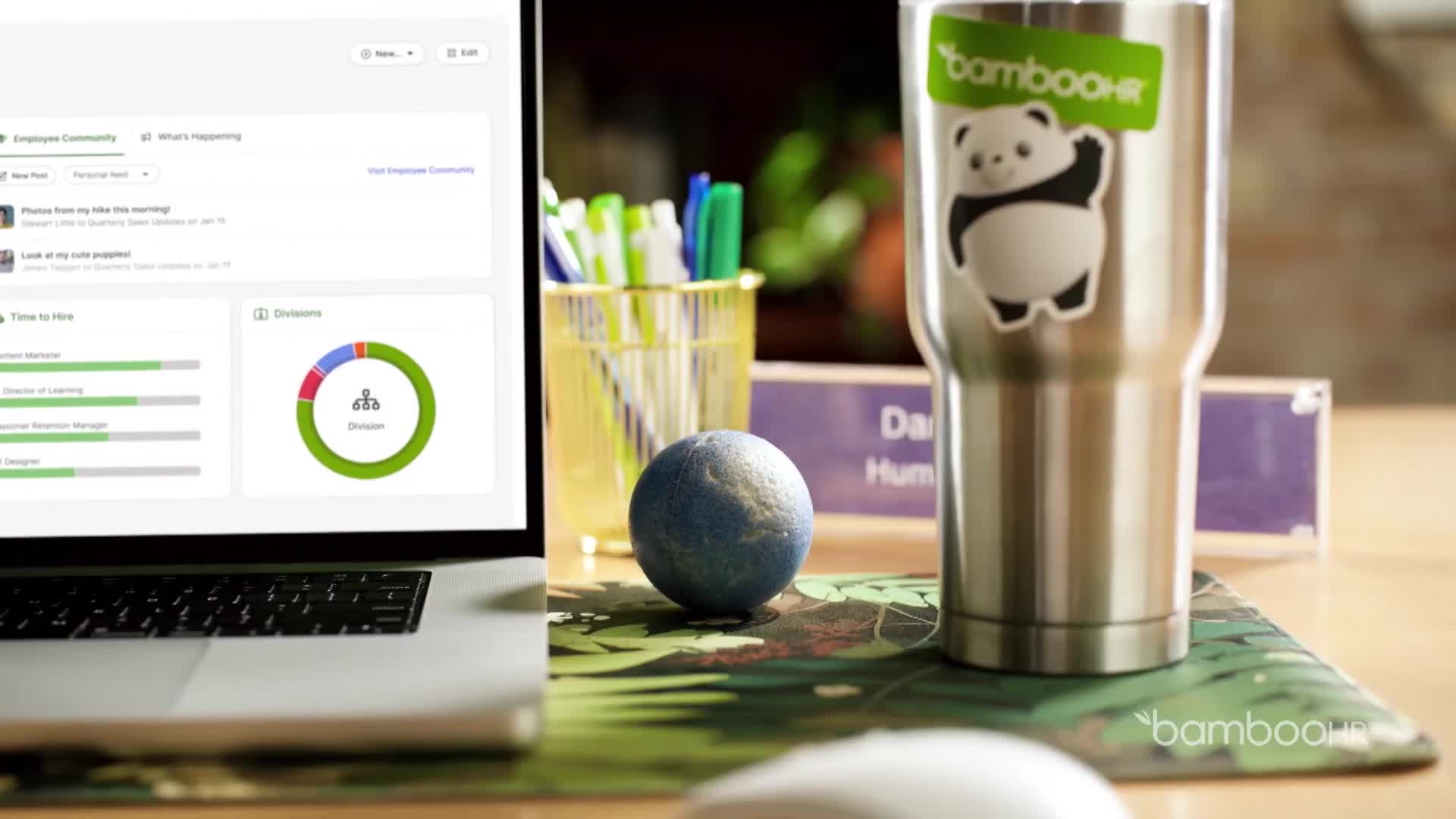 A laptop displays a dashboard with charts and graphs. Next to it, a metal tumbler with a panda sticker stands on a desk.