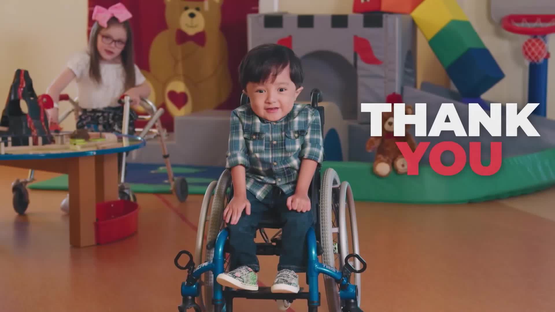 A young boy in a wheelchair smiles at the camera. A girl with a walker plays with toys at a table.