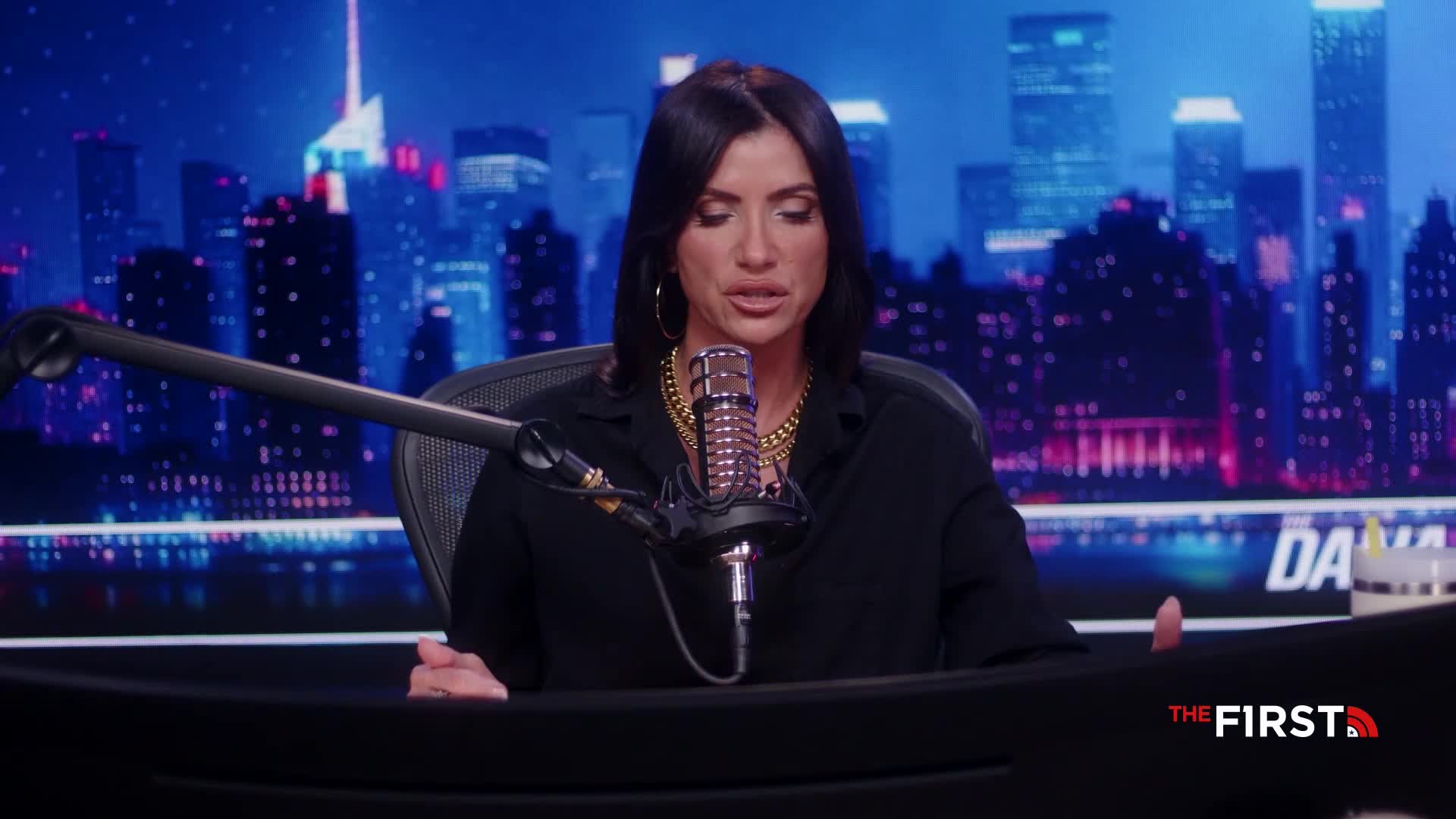 A woman with dark hair and a gold chain speaks into a microphone. Behind her, a cityscape glows under a blue light.