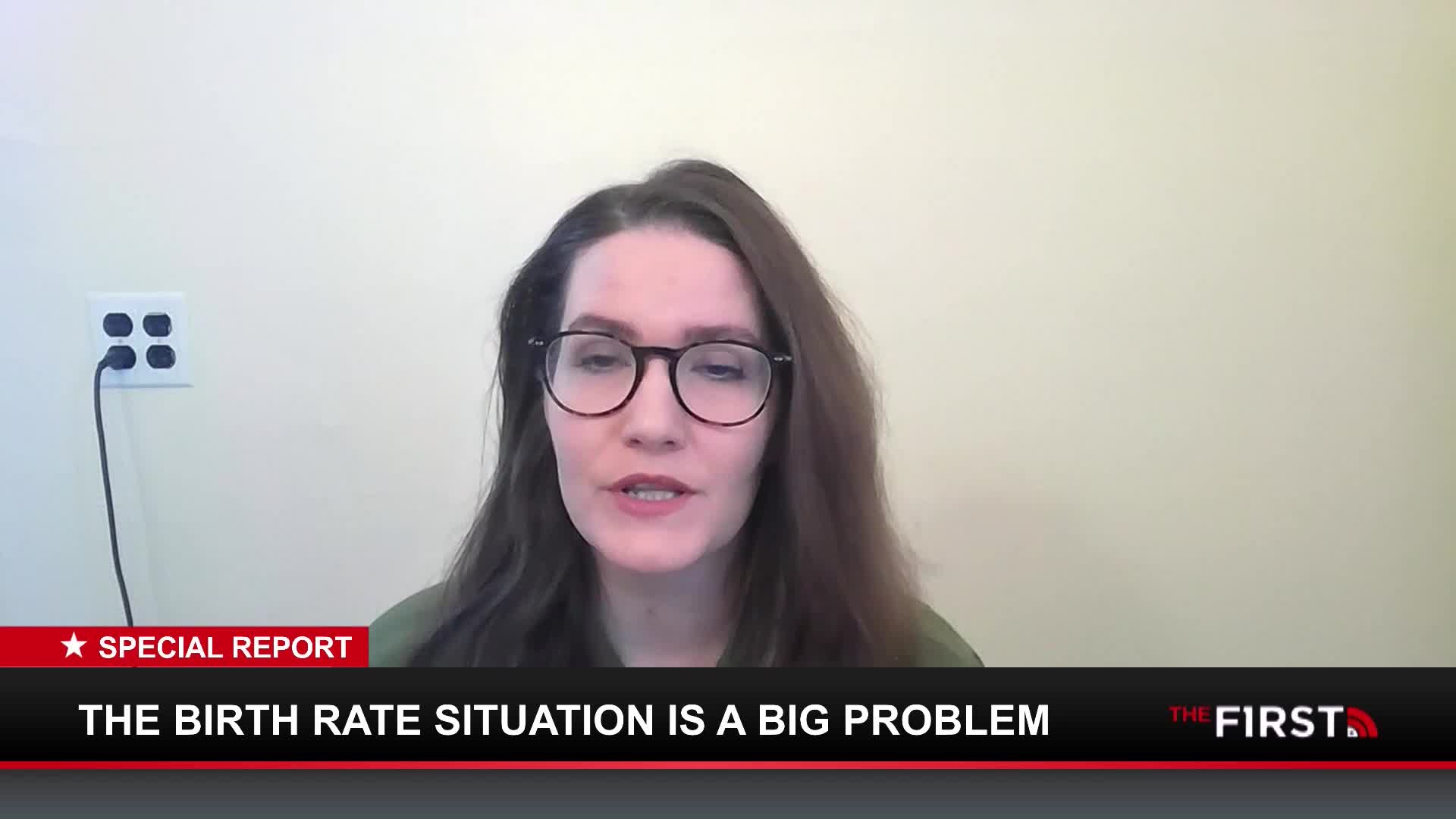 A woman with glasses speaks directly to the camera, her brown hair framing her face. A graphic overlay at the bottom of the screen reads "THE BIRTH RATE SITUATION IS A BIG PROBLEM" and "THE FIRST."