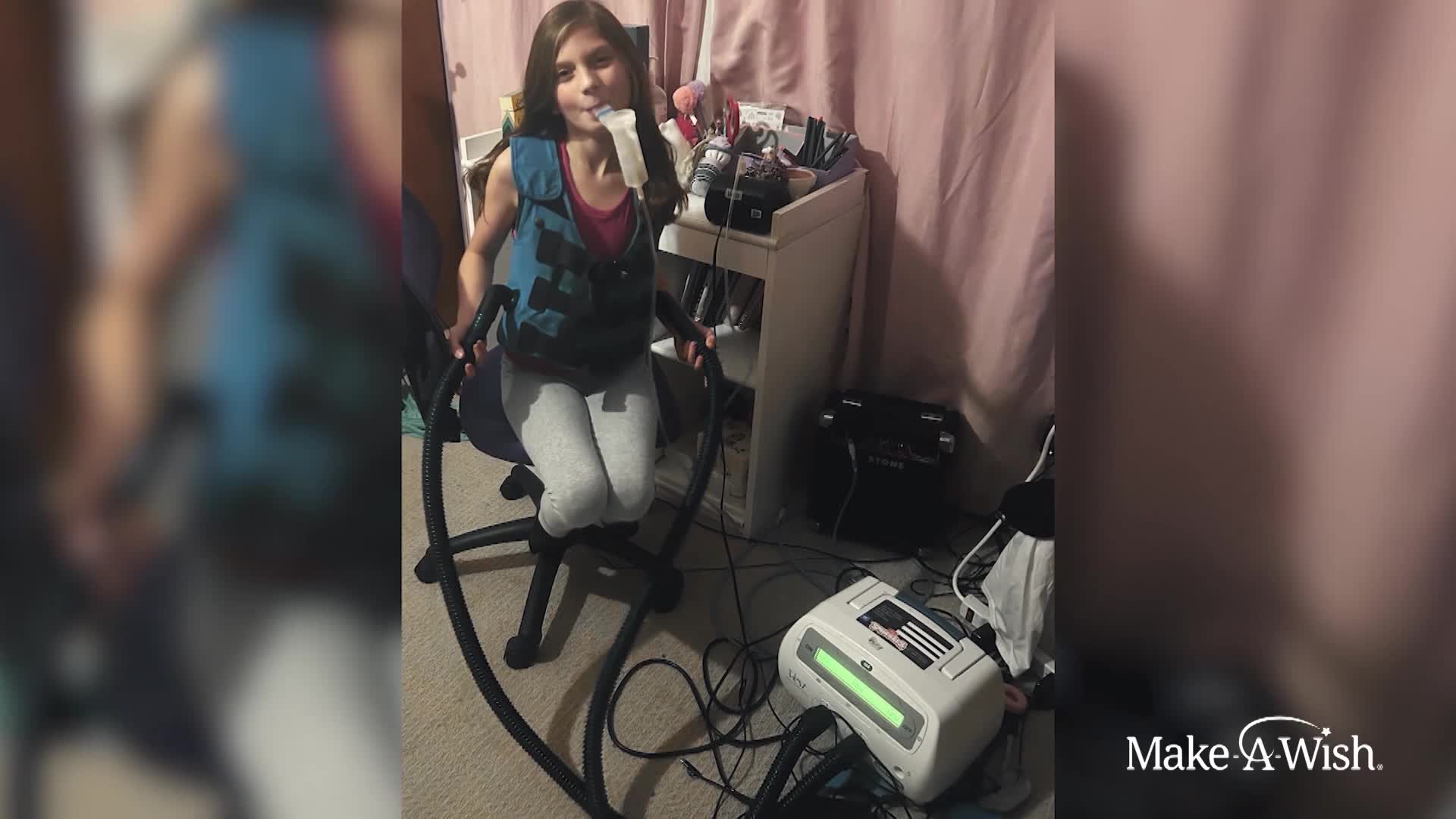 A young girl sits in a chair, holding a tube to her mouth. A chest vest and medical equipment surround her.