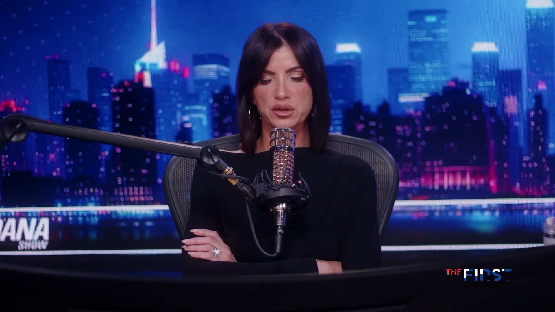 A woman with dark hair speaks into a microphone on a television set. Behind her, a cityscape glows with the lights of buildings.