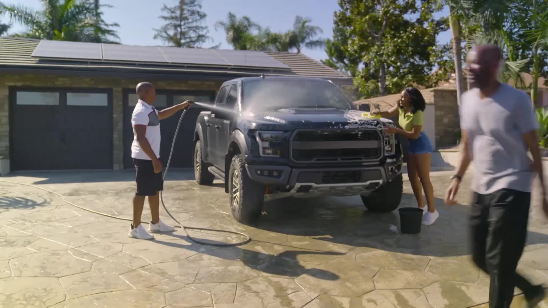 A man sprays water from a hose onto a black Ford Raptor while a woman soaps its hood. Another man walks past them, his back to the camera.