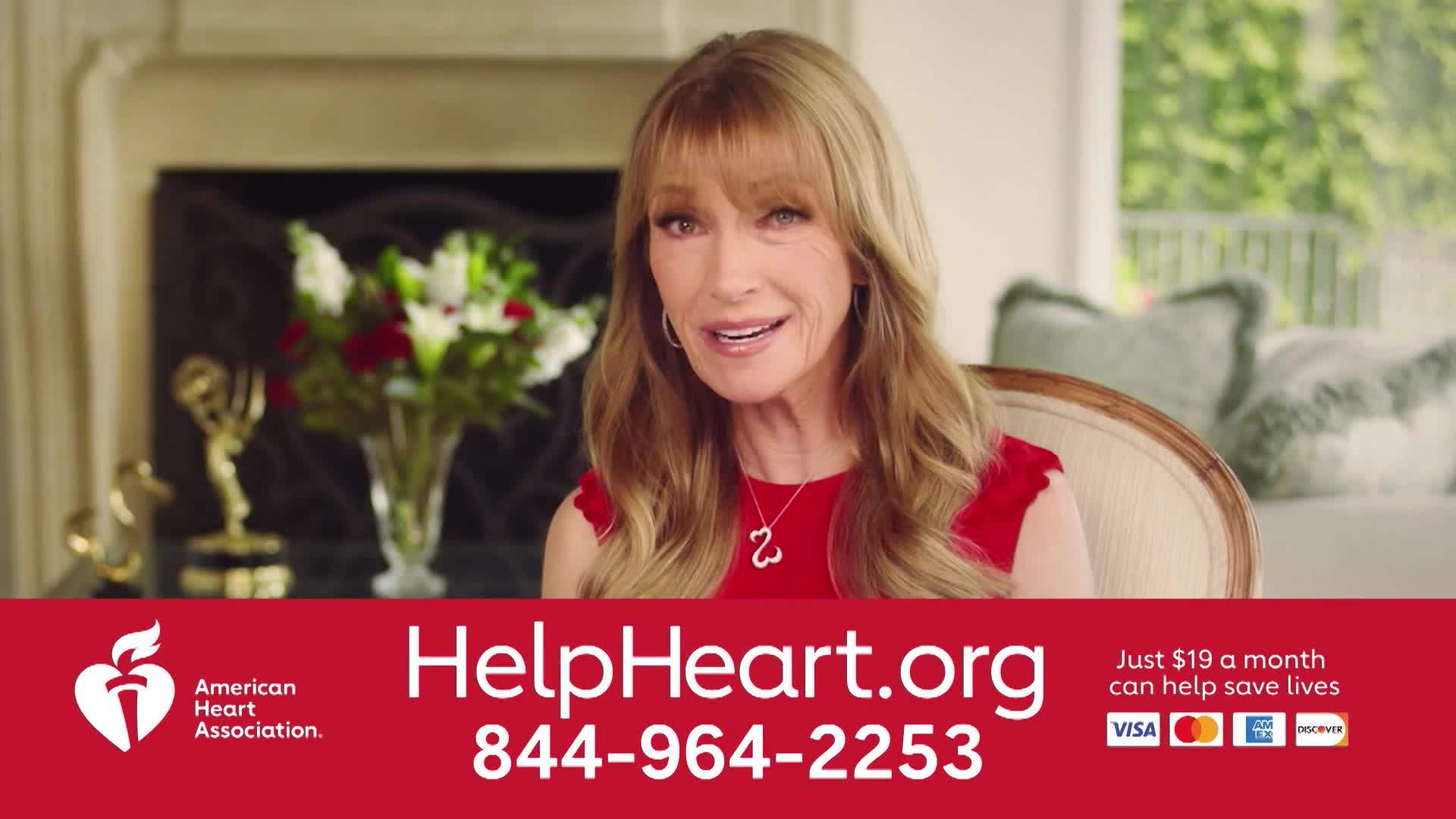 A woman with long blonde hair, wearing a red dress, speaks directly to the camera from a comfortable chair. Behind her, a fireplace and a vase of flowers are visible.