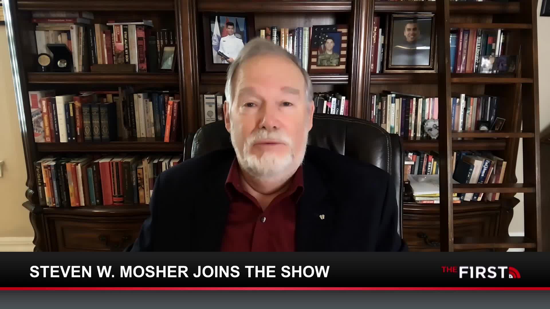 A man with a white beard sits in a leather chair, speaking directly to the camera. Behind him, bookshelves are filled with volumes, and framed photographs of military personnel are visible.