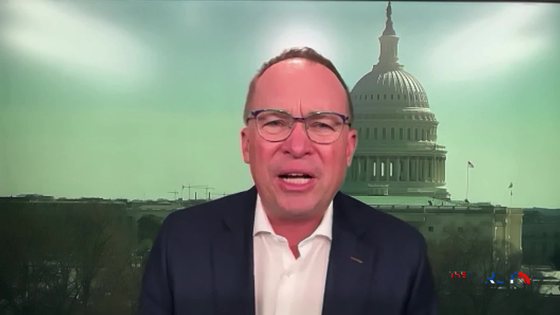 A man in a suit speaks directly to the camera, his expression animated. Behind him, the U.S. Capitol building stands under a hazy, pale green sky.