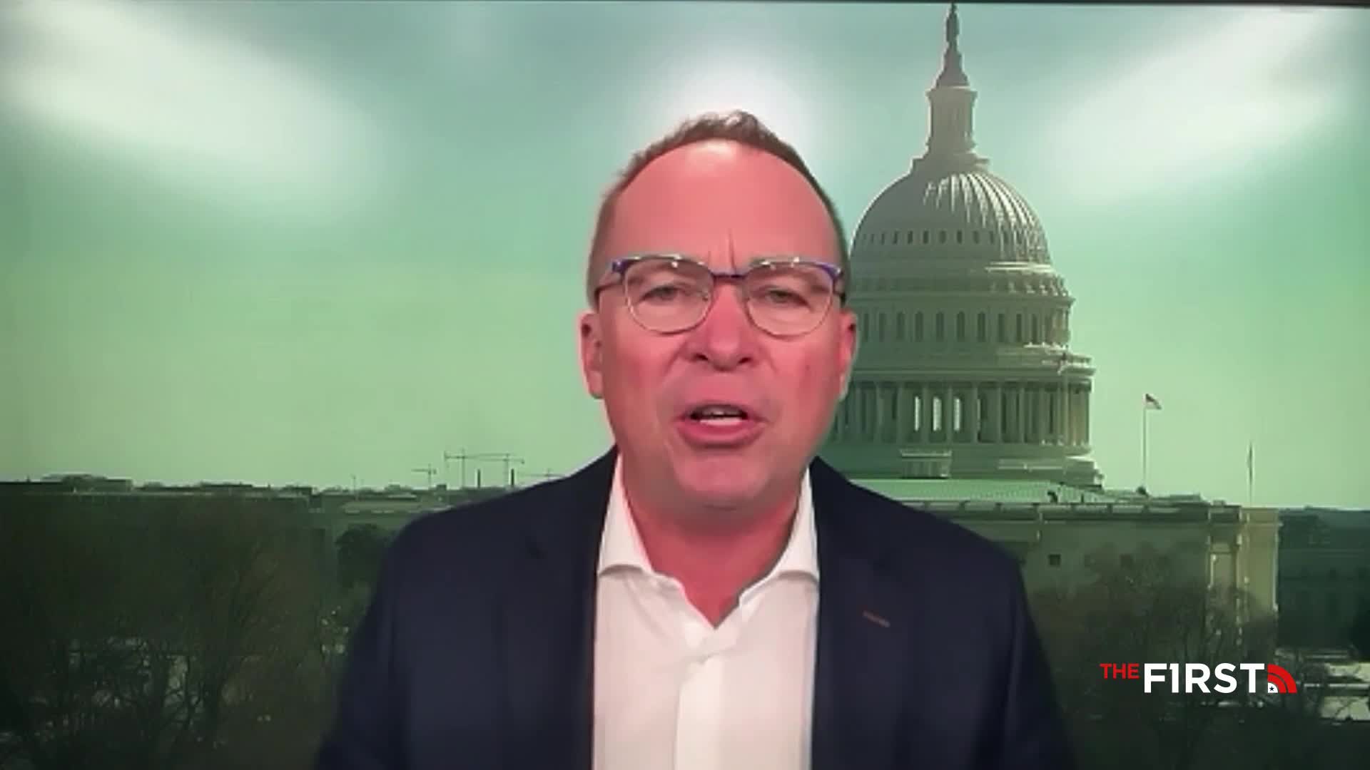 A man in a suit speaks directly to the camera, his face framed by a backdrop of the United States Capitol building. The logo for The First TV appears in the lower right corner.