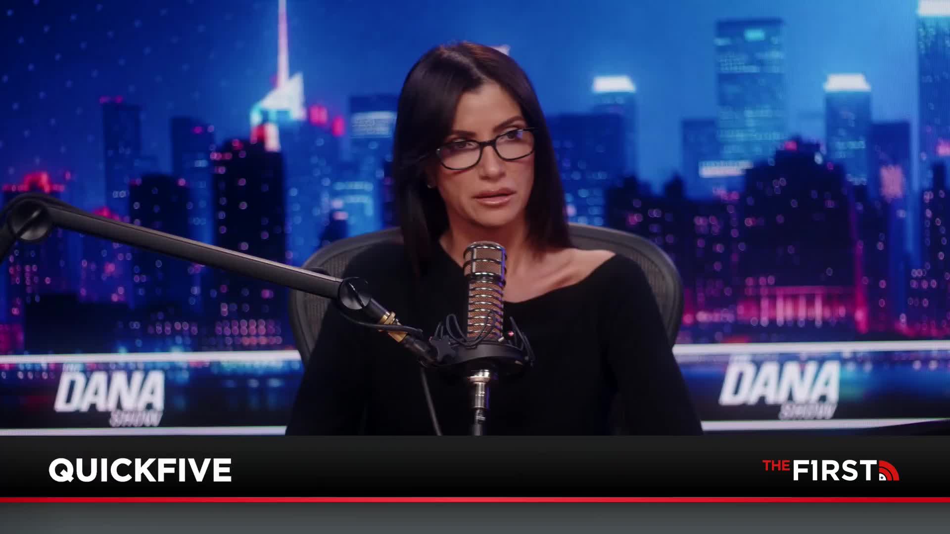 A woman with dark hair and glasses speaks into a microphone. The backdrop behind her displays a city skyline at night.