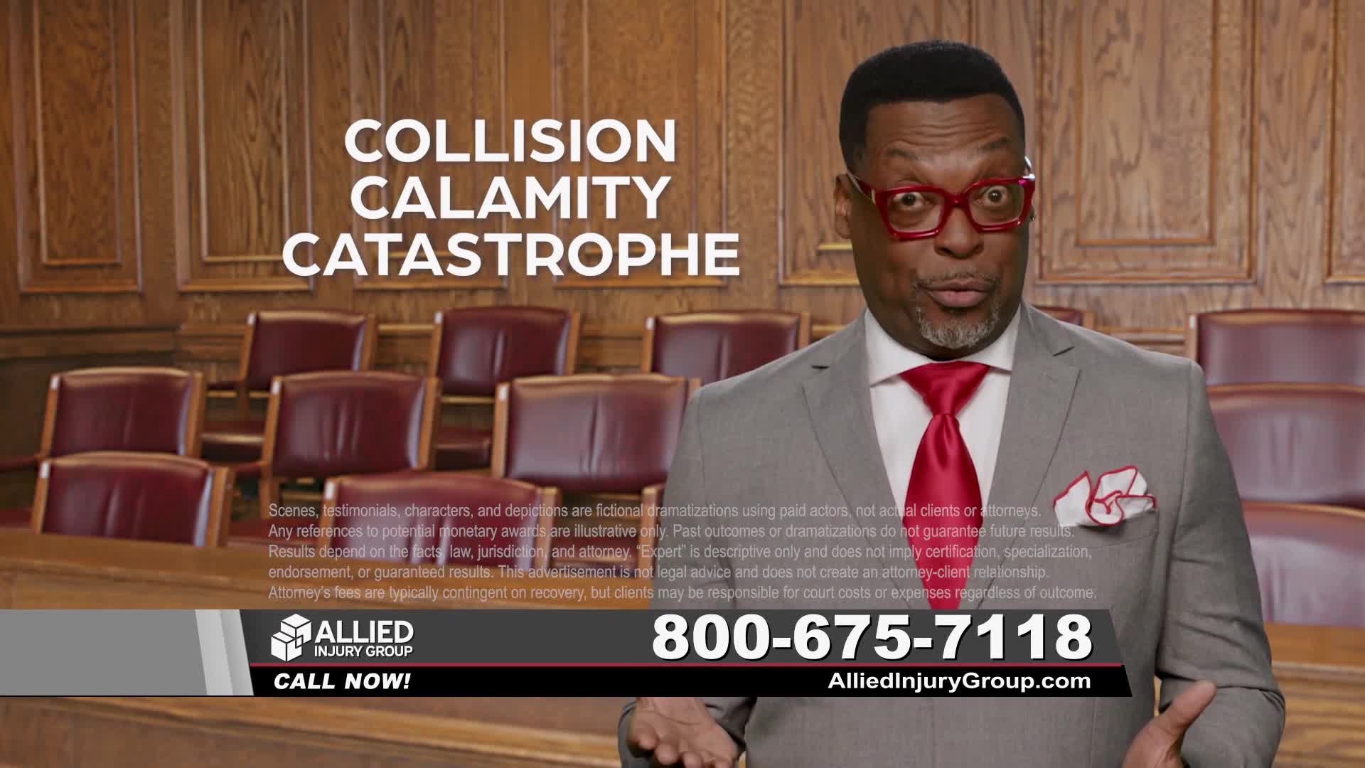 A man in a gray suit and red tie, wearing bright red glasses, gestures with his hands. He stands in front of rows of empty, dark red chairs in what looks like a courtroom.