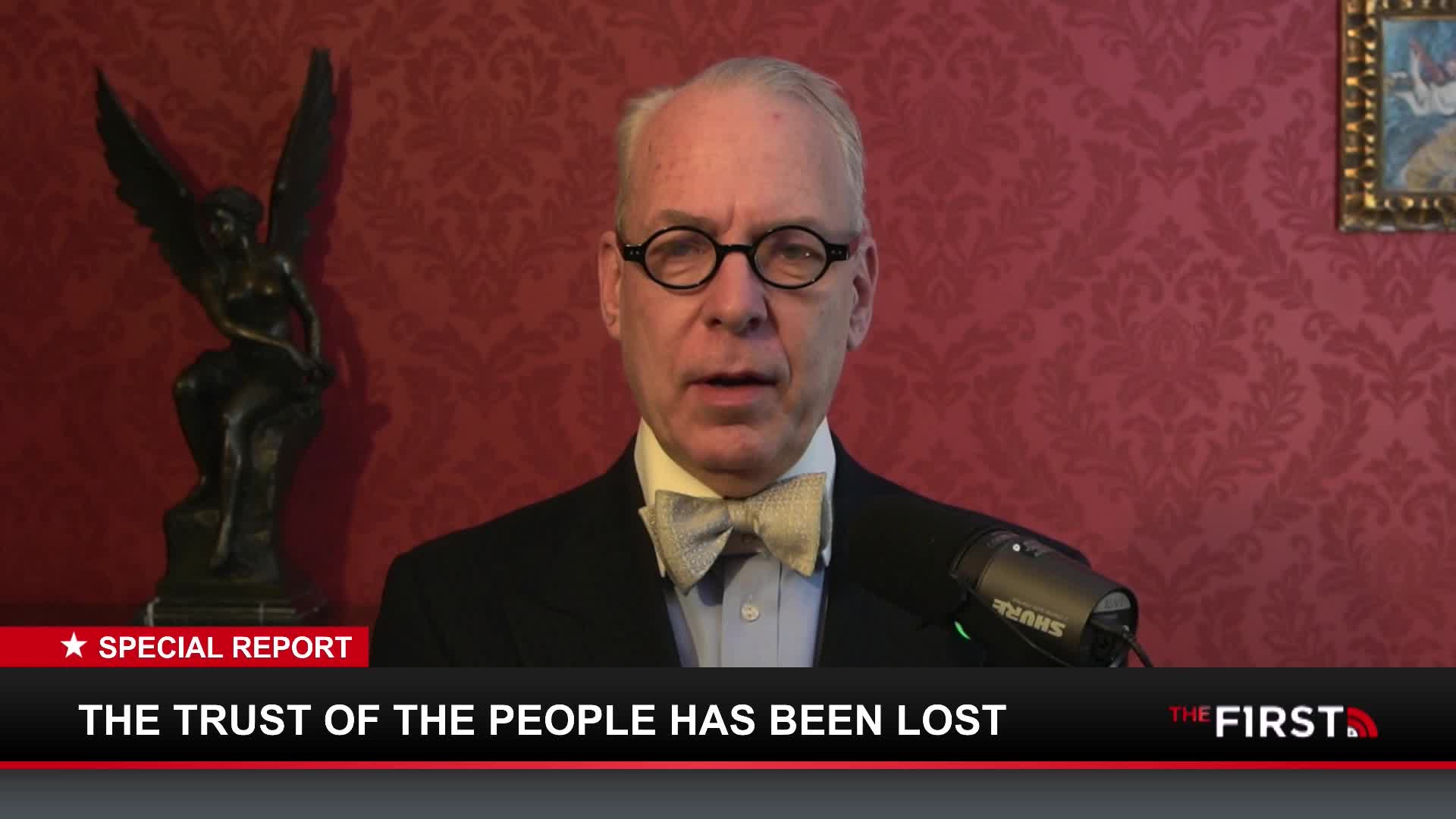 A man in a suit and bow tie speaks into a microphone. A graphic below him reads "THE TRUST OF THE PEOPLE HAS BEEN LOST."