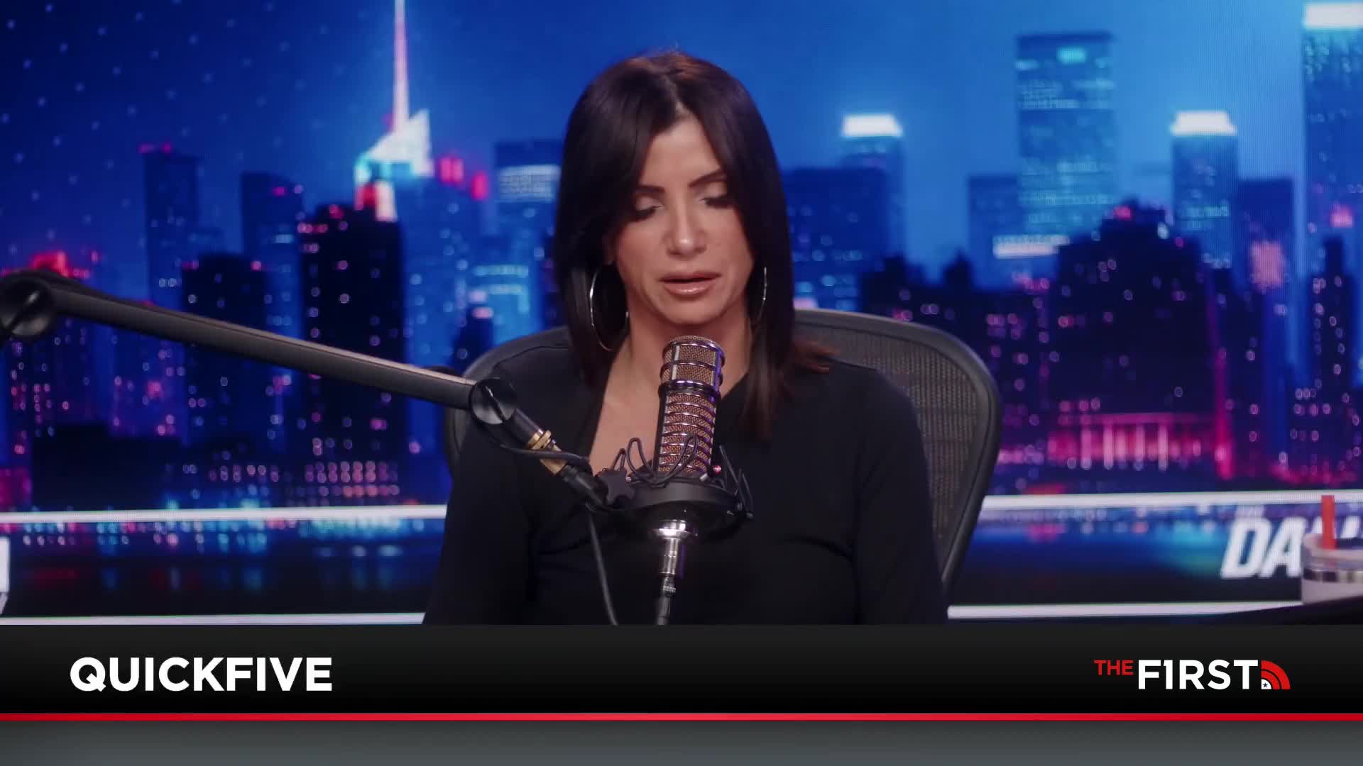 A woman with dark hair sits at a desk, speaking into a microphone. Behind her, a cityscape glows with neon lights.