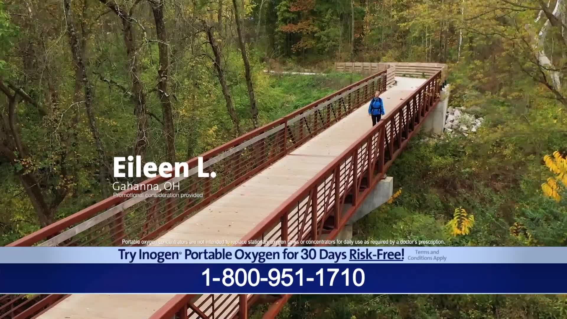 A woman in a blue jacket walks across a rust-colored bridge. She's heading away from the camera, into a wooded area with autumn leaves.