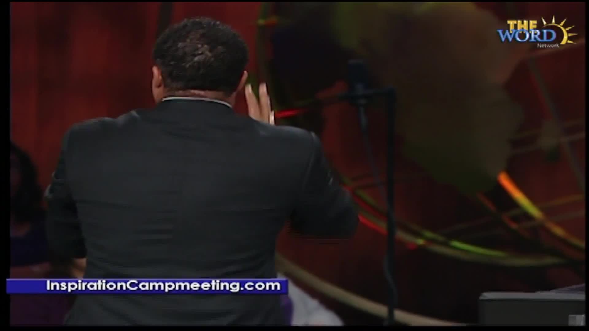 A man in a black suit raises his hands, fingers spread, as if in prayer or blessing. The logo for The Word Network is visible in the corner.
