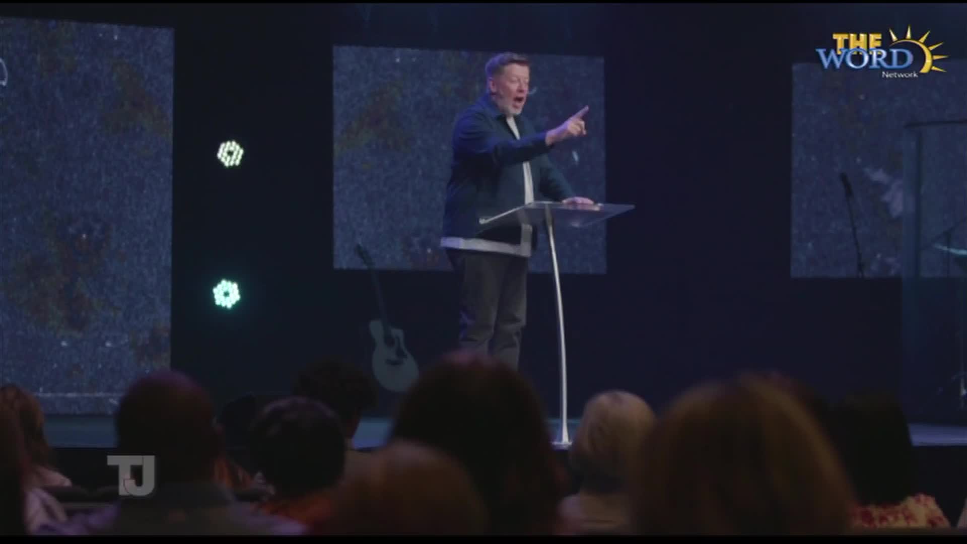 A man in a dark jacket gestures emphatically, pointing his finger as he speaks from behind a clear podium. The Word Network logo is visible in the corner, and the audience is focused on the stage.

