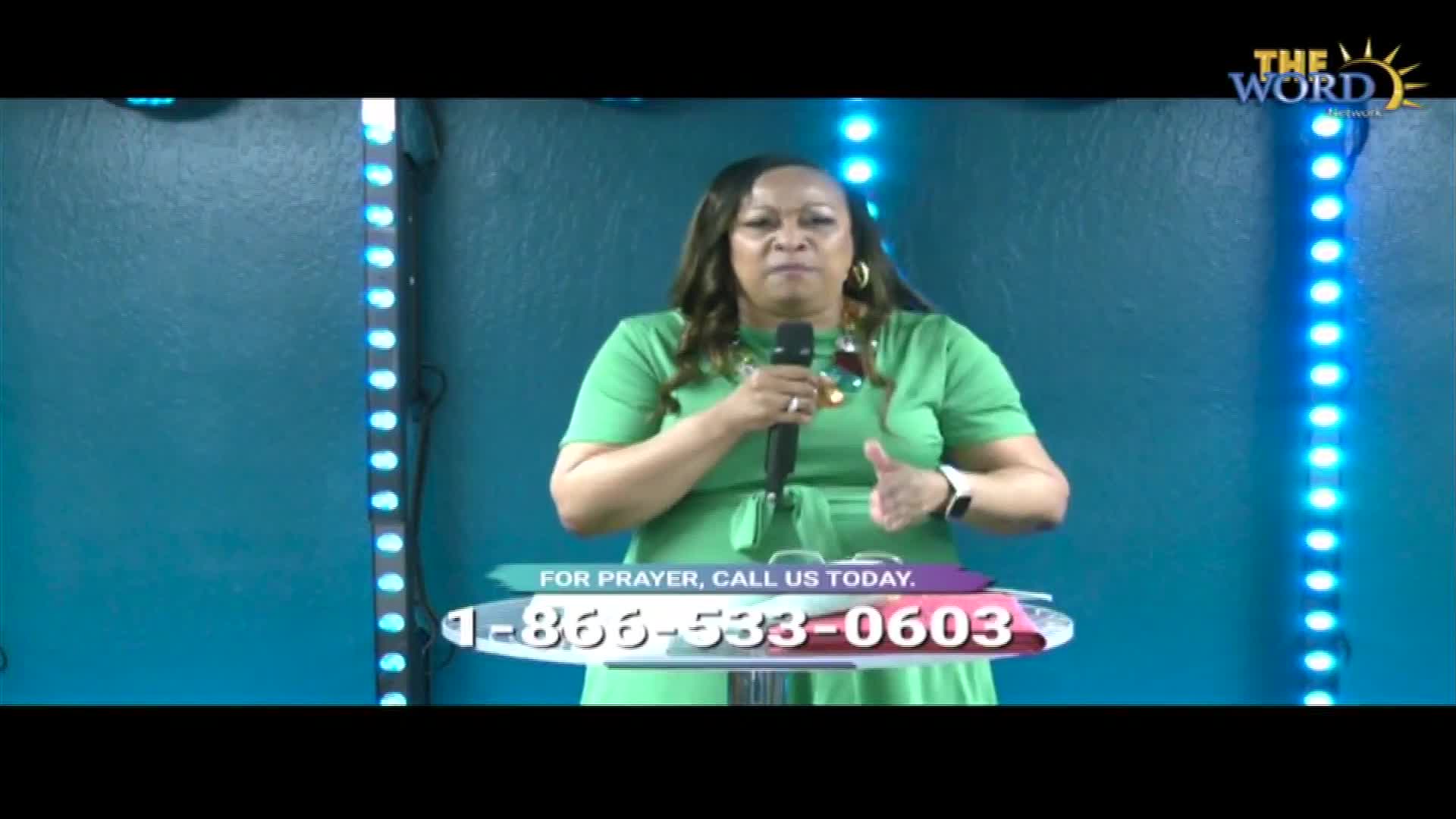 A woman in a green dress gestures with one hand while holding a microphone. The Word Network logo is visible, and a phone number for prayer is displayed at the bottom.
