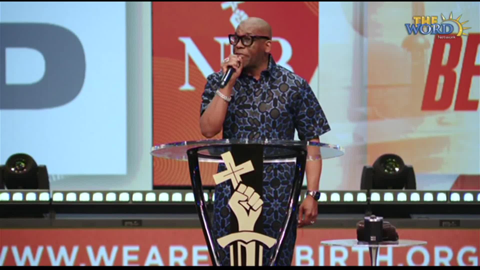 A man in a patterned shirt stands at a transparent podium, speaking into a microphone. Behind him, the backdrop displays "The Word Network" logo and large letters.
