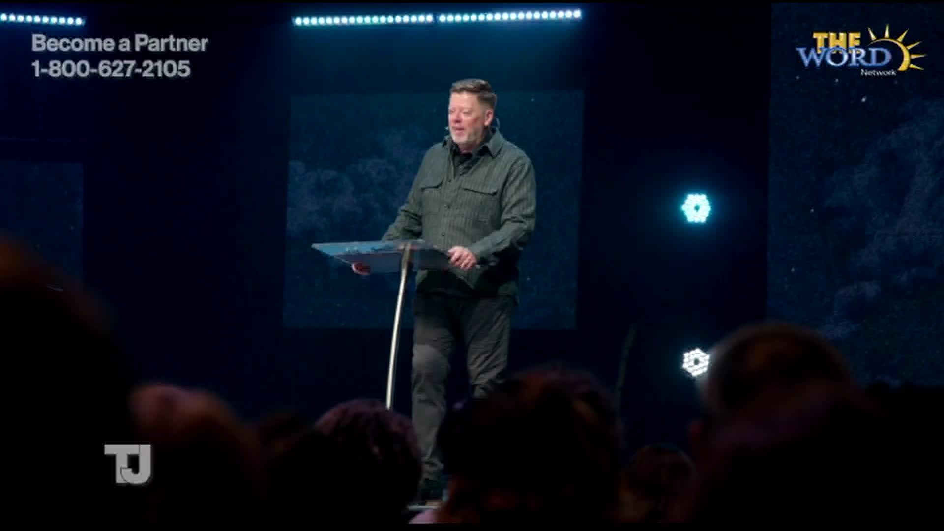 A man in a green shirt stands at a clear podium, speaking to an unseen audience. Behind him, a dark blue backdrop features the logo for The Word Network and some floating, geometric lights.
