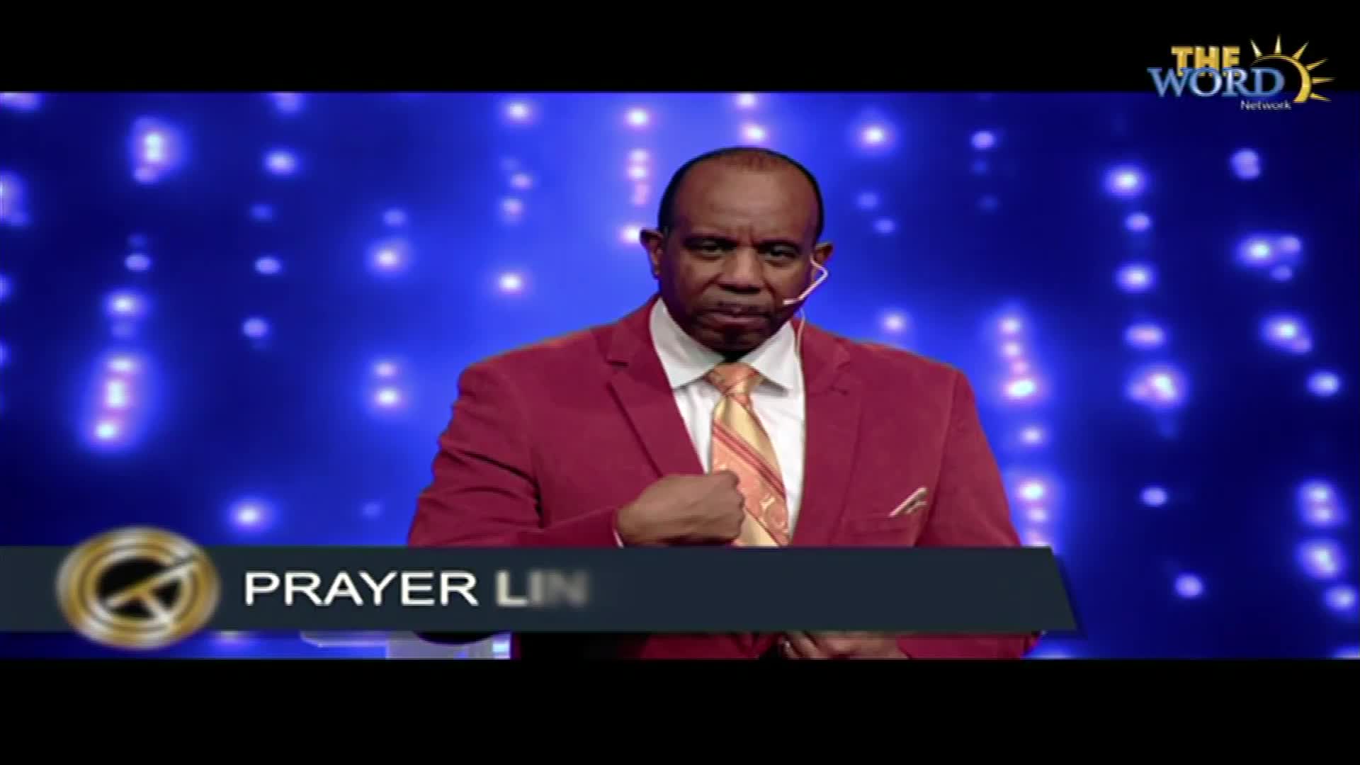 A man in a red suit stands center stage, his hand resting on his chest. The background is a deep blue, dotted with bright lights.
