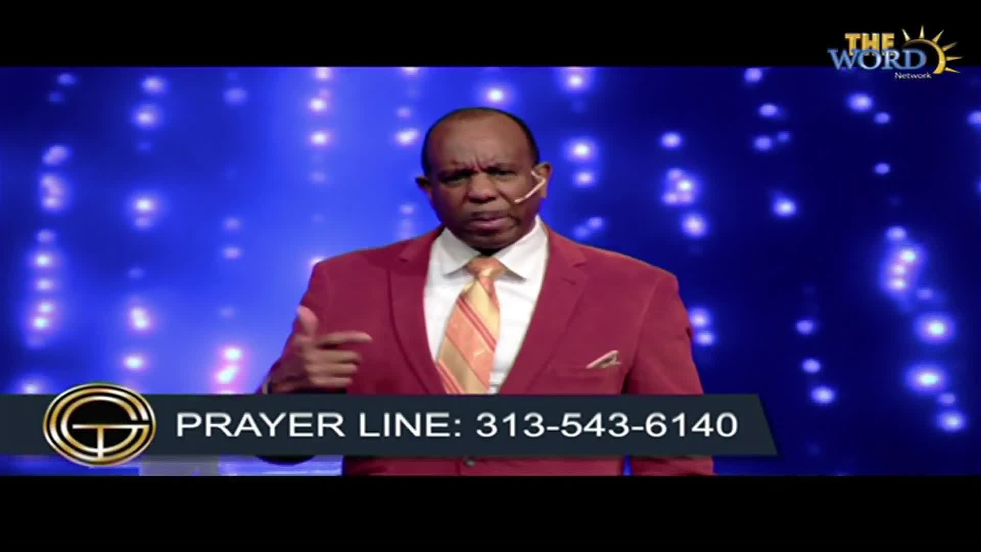 A man in a red suit gestures with his left hand, speaking on a stage with a blue backdrop illuminated by small lights. Below, a graphic on the screen displays a phone number for a prayer line.
