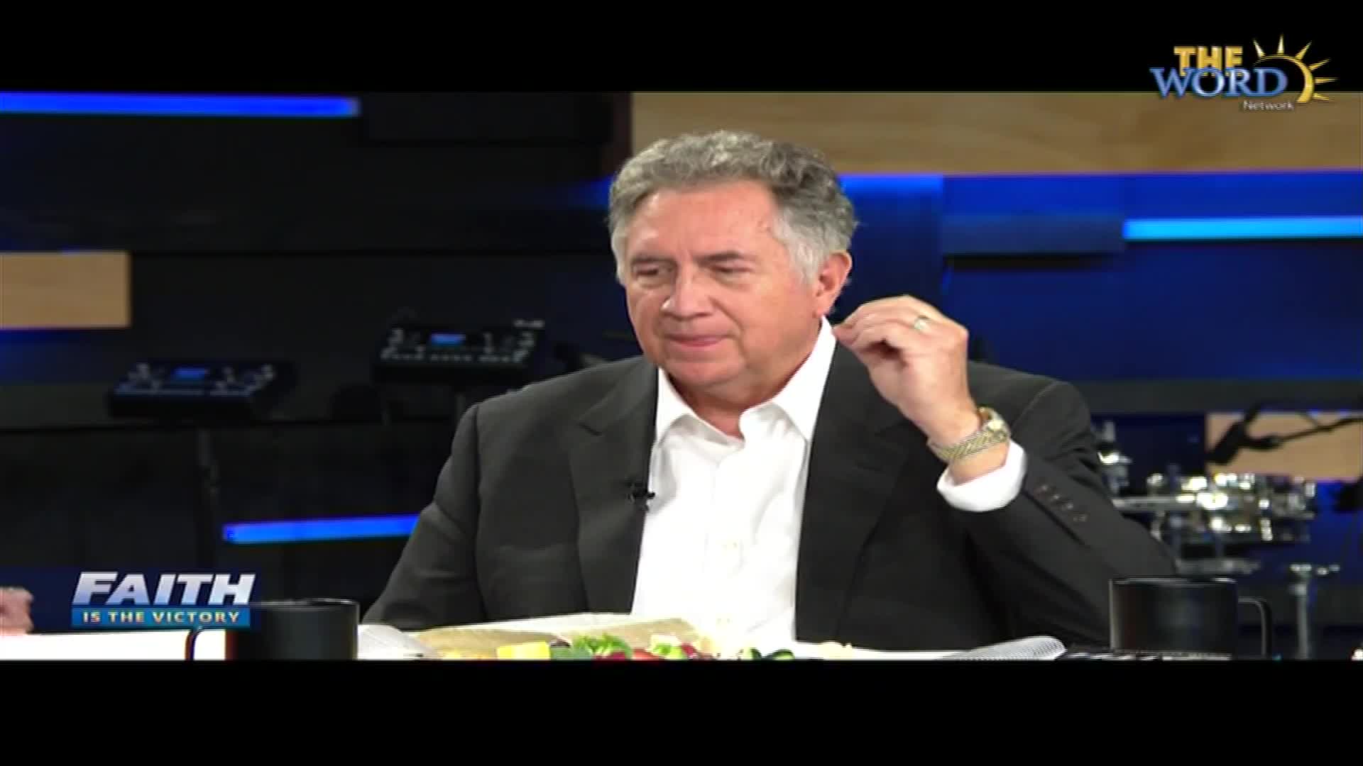 A man in a suit, seated at a table, adjusts his ear. The Word Network logo is visible above him.
