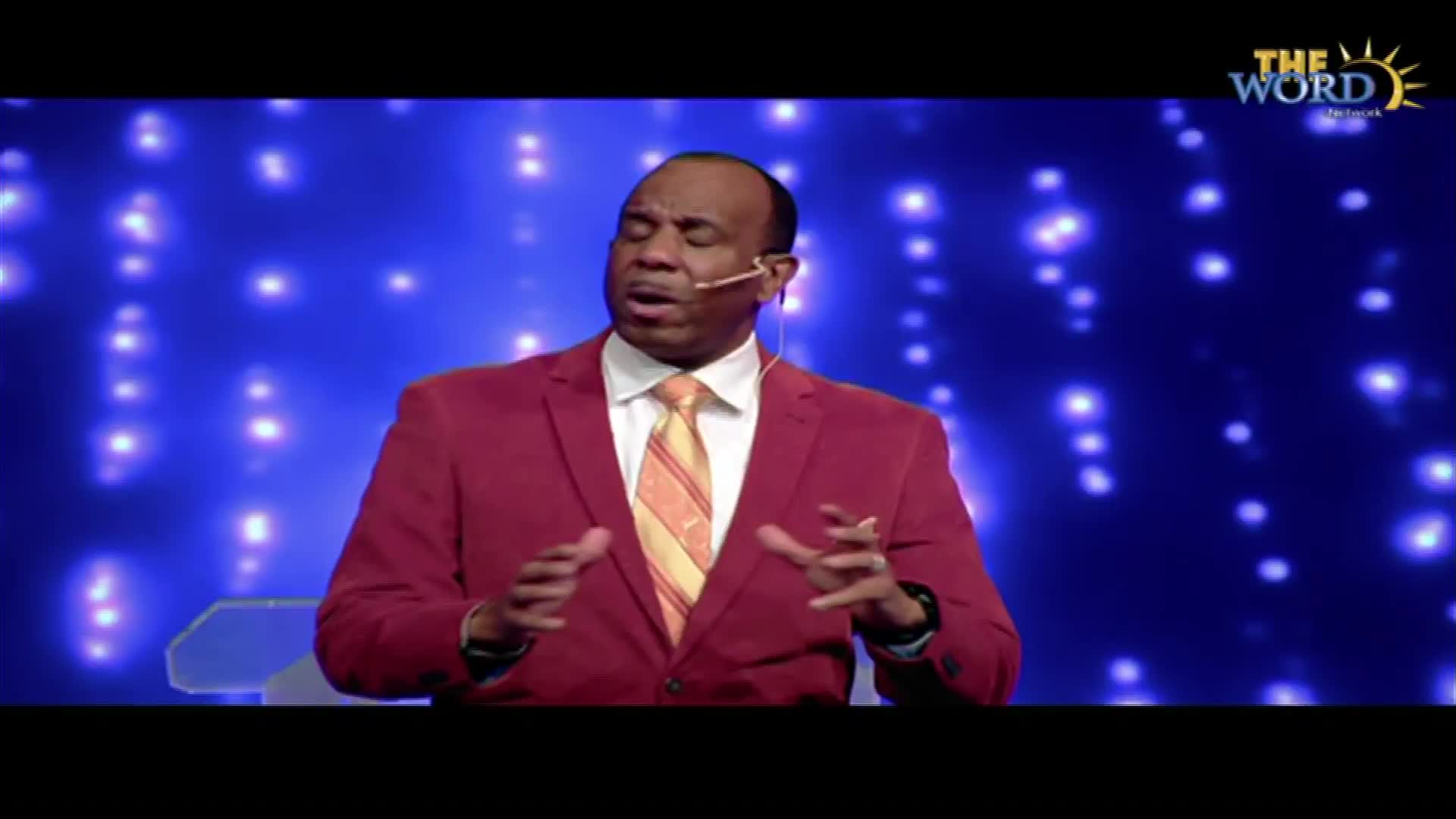 A man in a red suit, white shirt, and patterned tie speaks into a microphone. His hands move as he gestures toward the blue backdrop dotted with lights.
