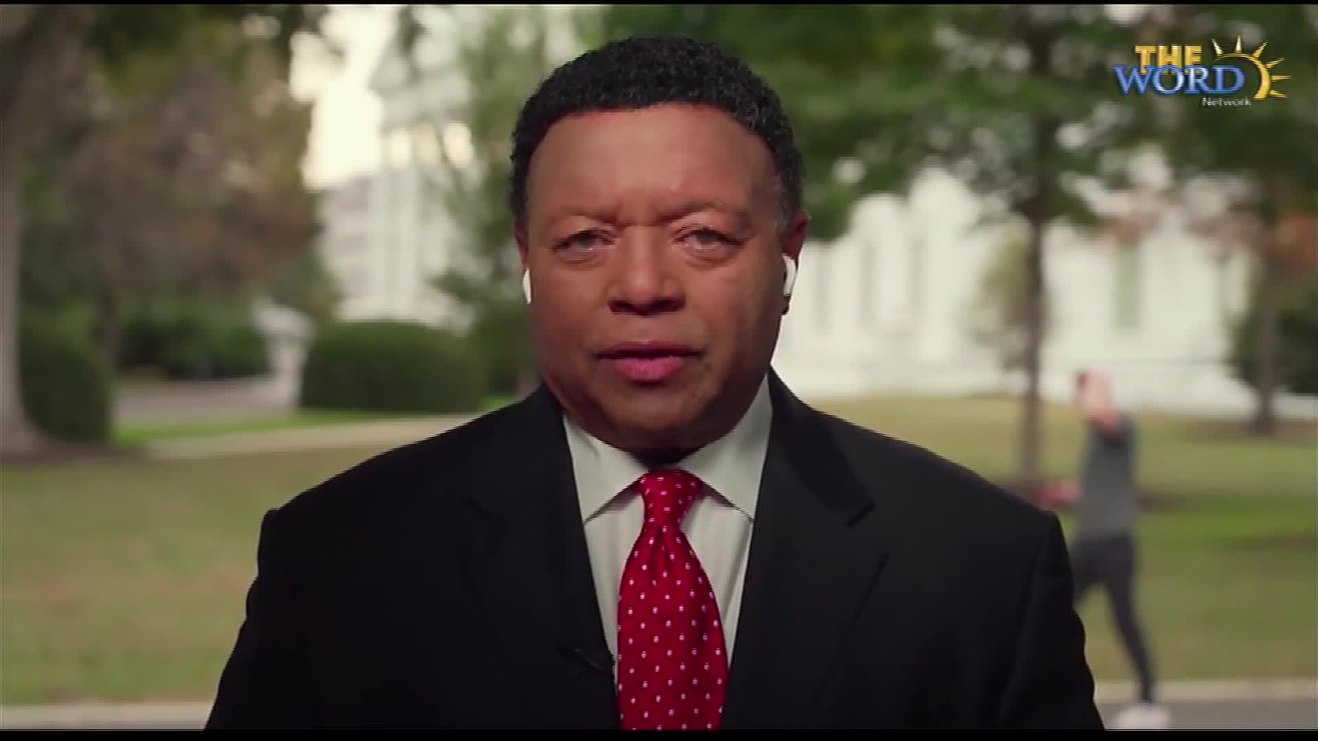 A man in a suit and red tie, wearing earbuds, looks directly at the camera. The Word Network logo is visible, and the backdrop suggests a location in the United States with a building and trees.
