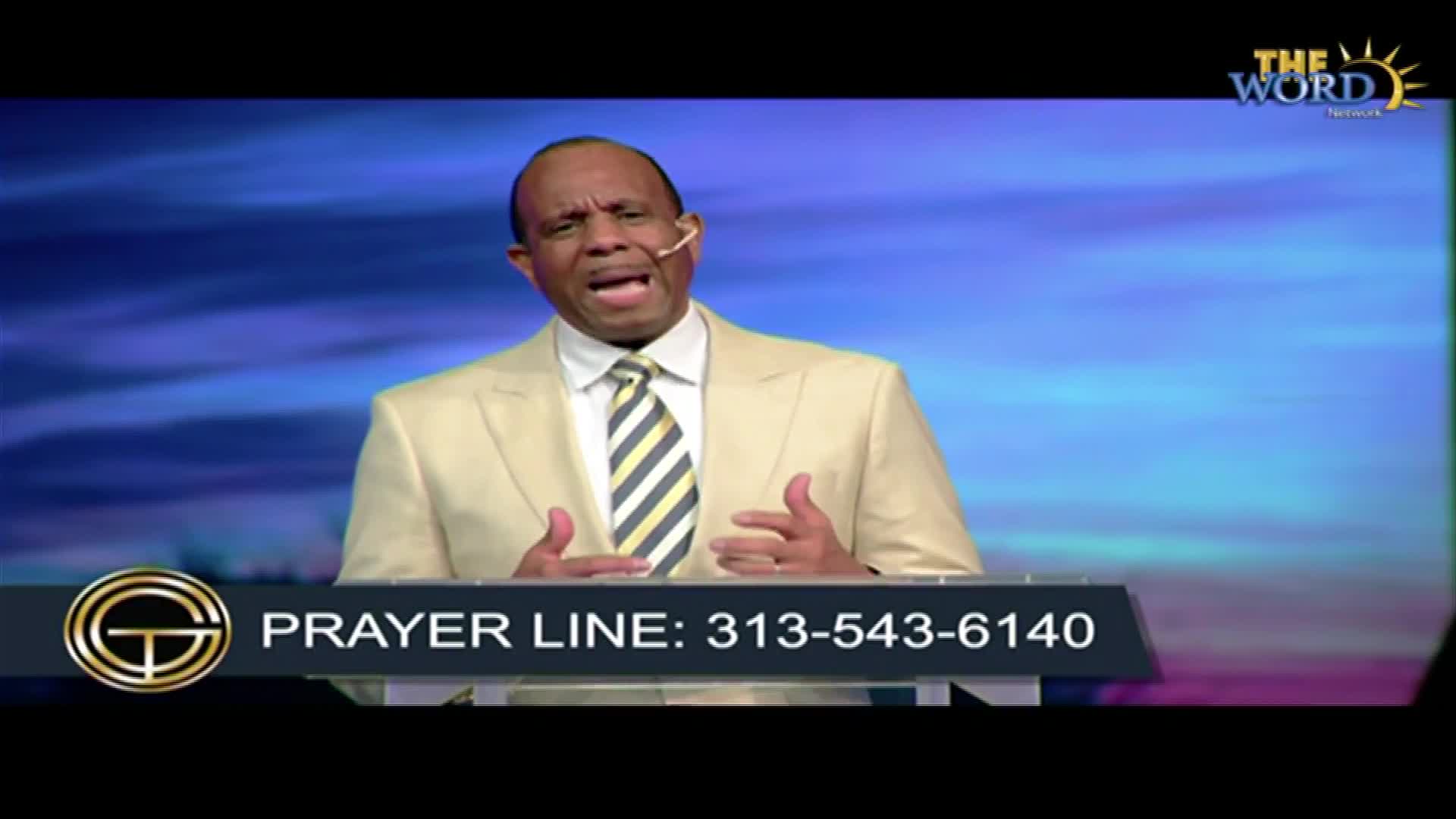A man in a light-colored suit speaks passionately, his hands gesturing as he addresses the camera. Below, a phone number for a prayer line is displayed on the screen.
