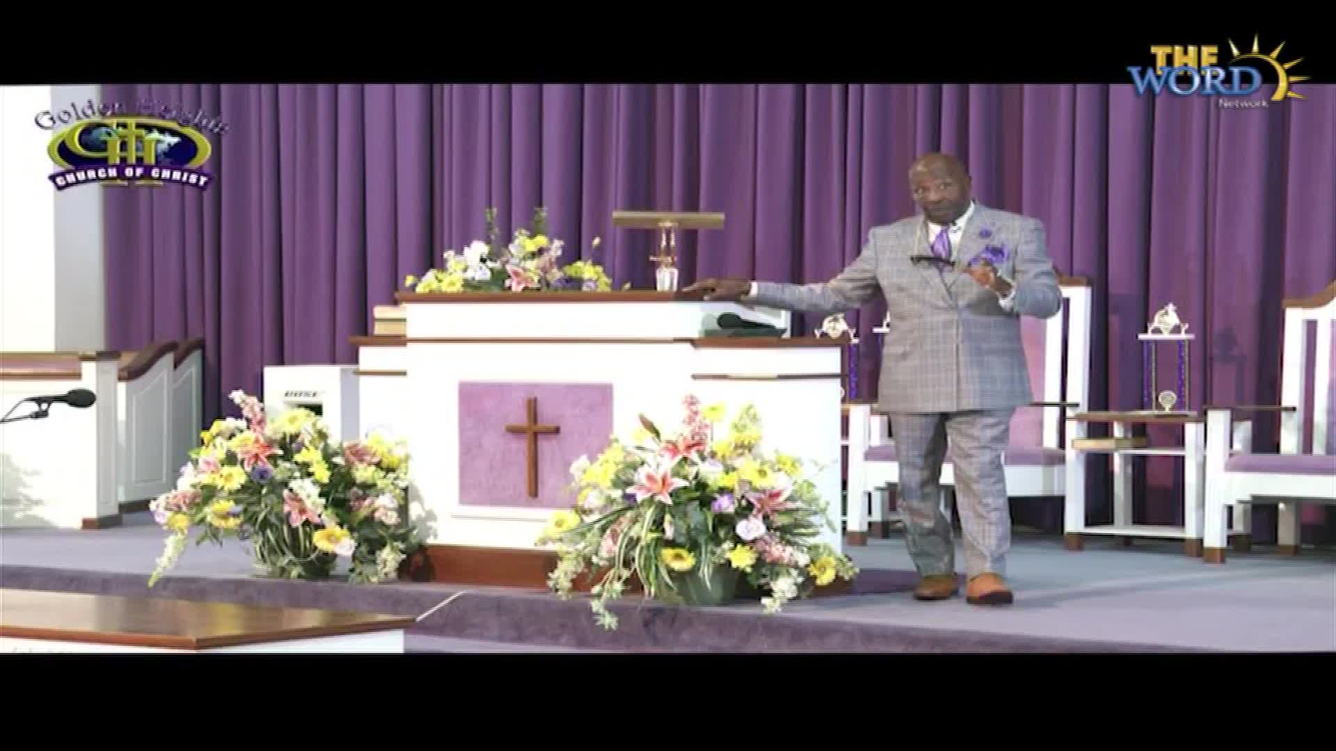 A man in a gray suit gestures toward the altar as he speaks, his right hand resting on the edge. The scene is set in a church, with the logo for The Word Network visible in the corner.
