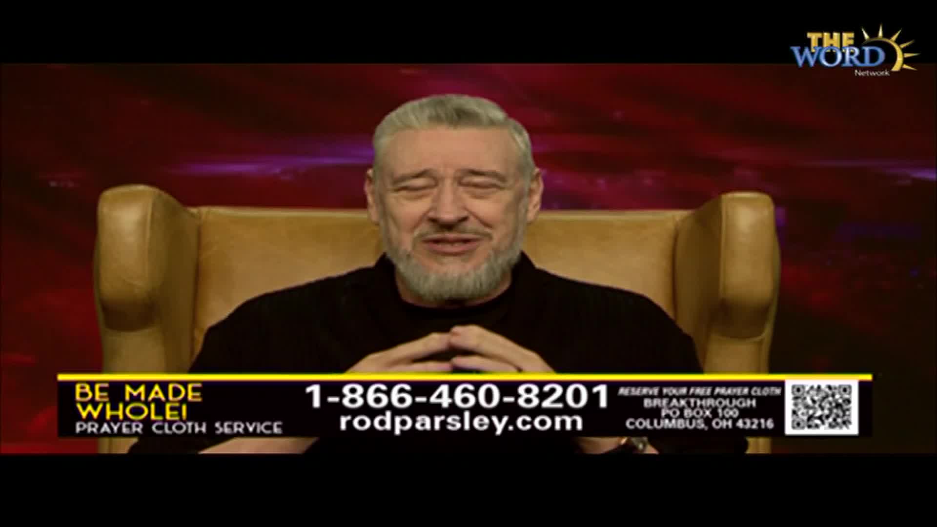 A man with a silver beard sits in a tan armchair, speaking on The Word Network. Below him, a banner advertises a prayer cloth service, complete with a phone number and website.
