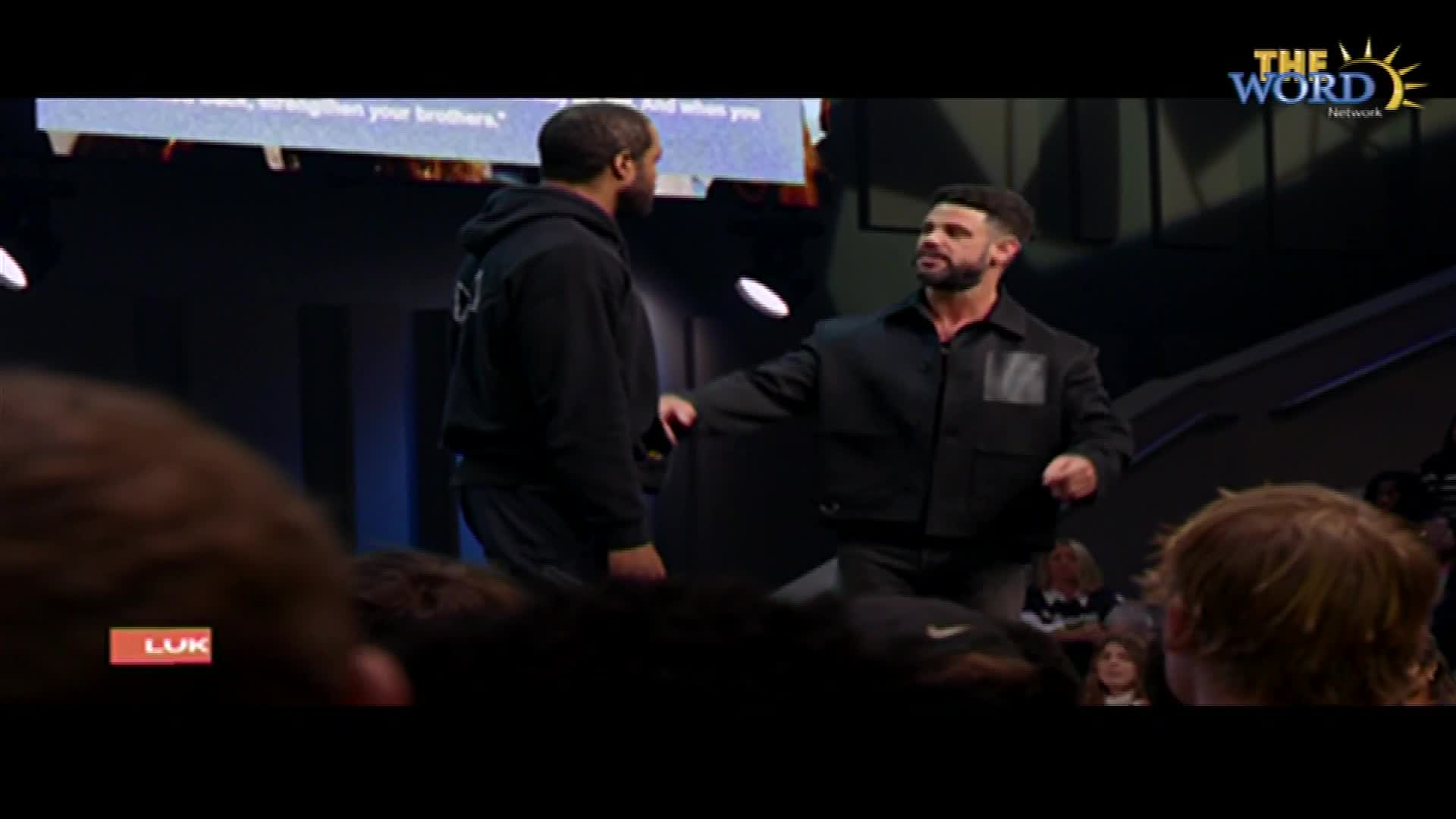 A man in a black jacket gestures toward another man wearing a hoodie. The Word Network logo sits in the upper right corner.
