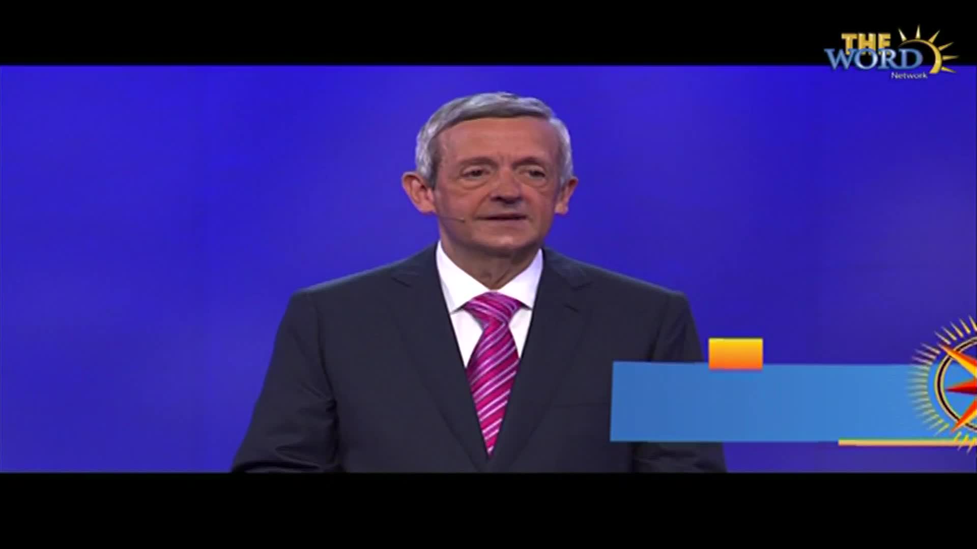 A man in a suit and tie is speaking on a stage, with the logo for The Word Network visible in the corner. The background is a solid blue, and a graphic is displayed below him.
