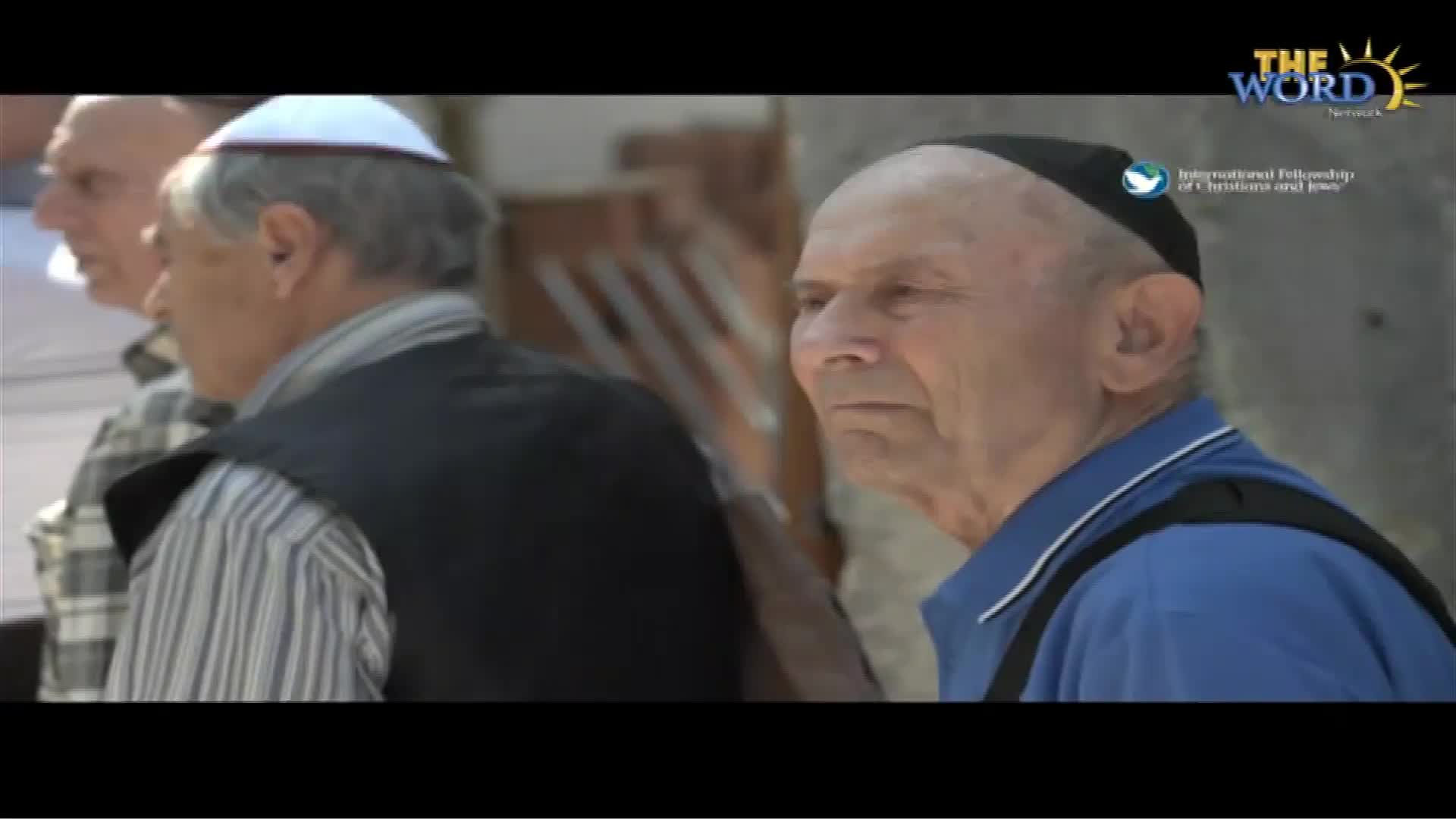 A man in a blue shirt and black kippah looks off-camera, his expression serious. Two other men, one in a vest and a white kippah, stand nearby.
