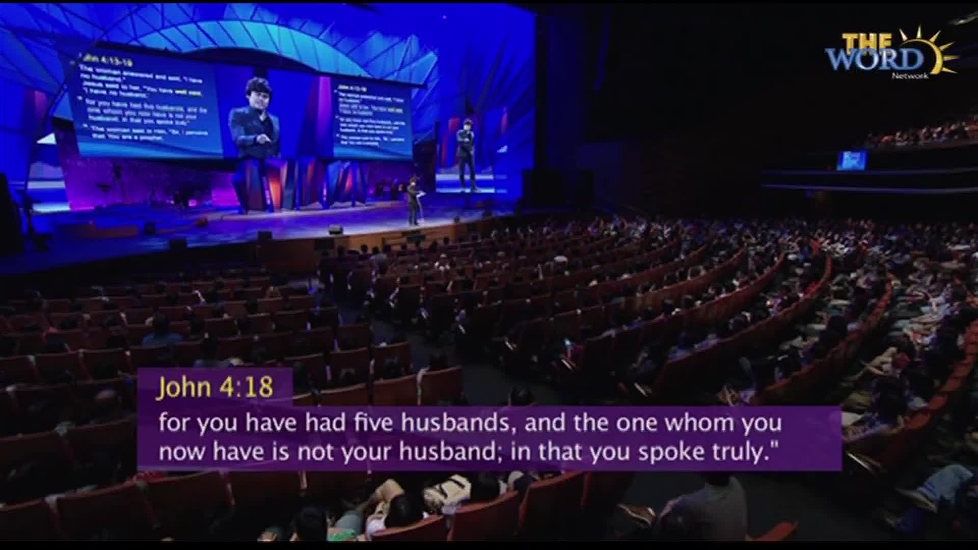 A man stands on a stage, speaking into a microphone with text from John 4:18 displayed below. The Word Network logo is visible, and the audience fills the curved seating.
