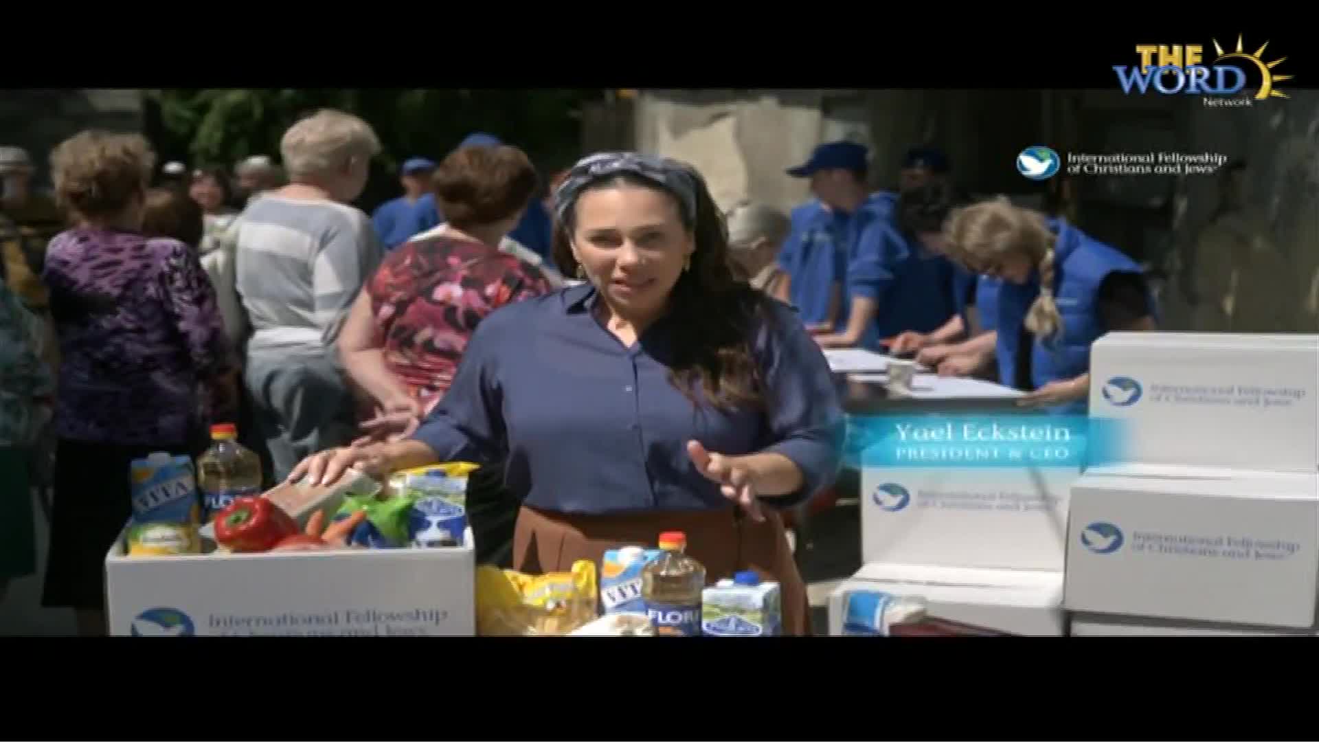 A woman is speaking, gesturing toward boxes of food and a crowd of people. Behind her, volunteers in blue vests are at a table, likely organizing the distribution.
