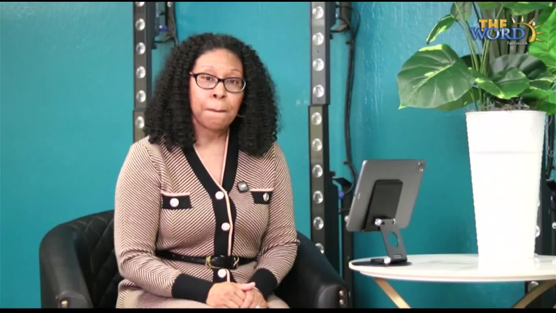 A woman in a patterned sweater sits in a chair, looking directly at the camera. She's in a studio setting, likely for a broadcast on The Word Network.
