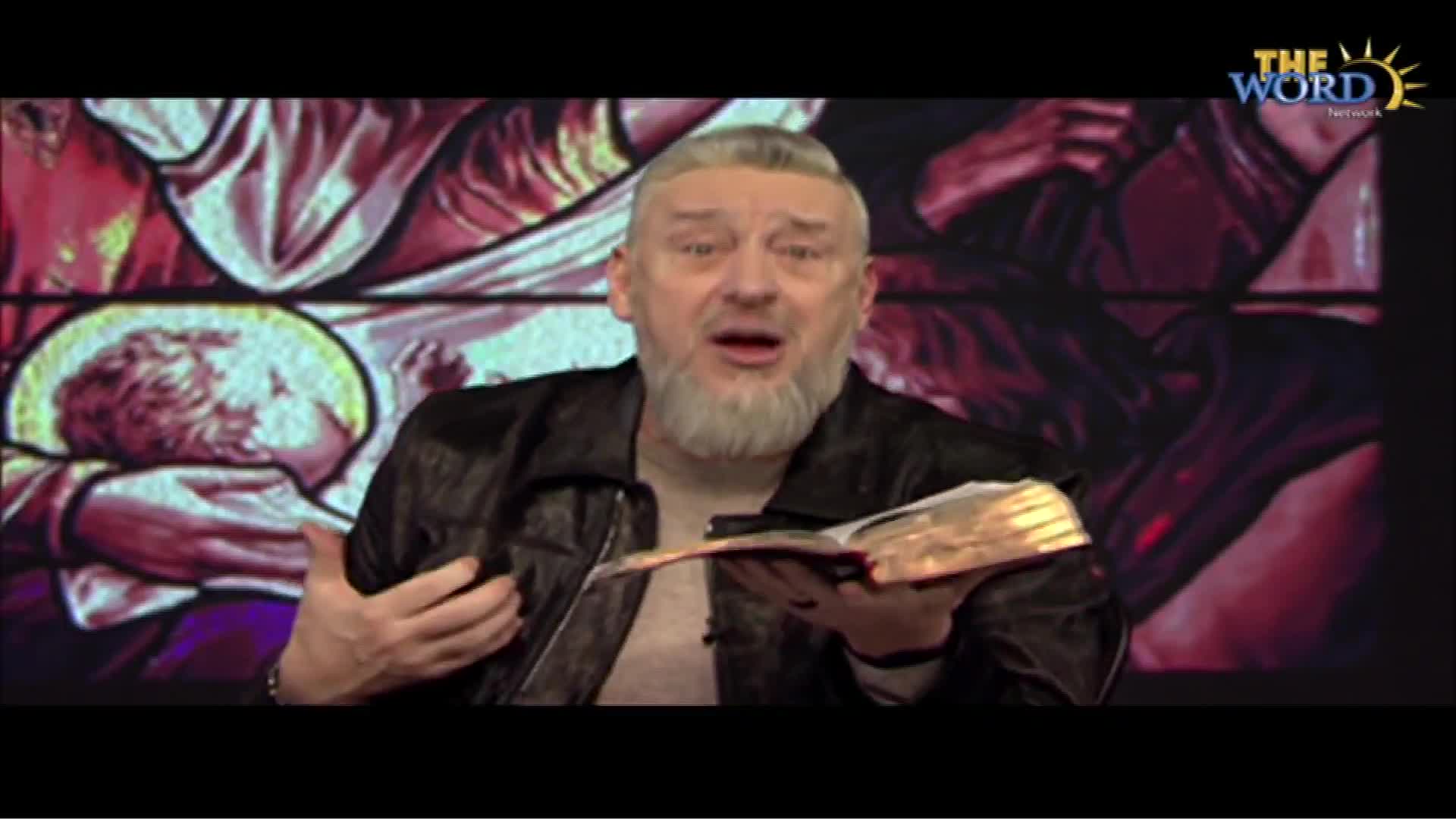 A man with a white beard and a leather jacket gestures emphatically while holding an open book. Behind him, a stained-glass window provides a backdrop for what appears to be a broadcast on The Word Network.
