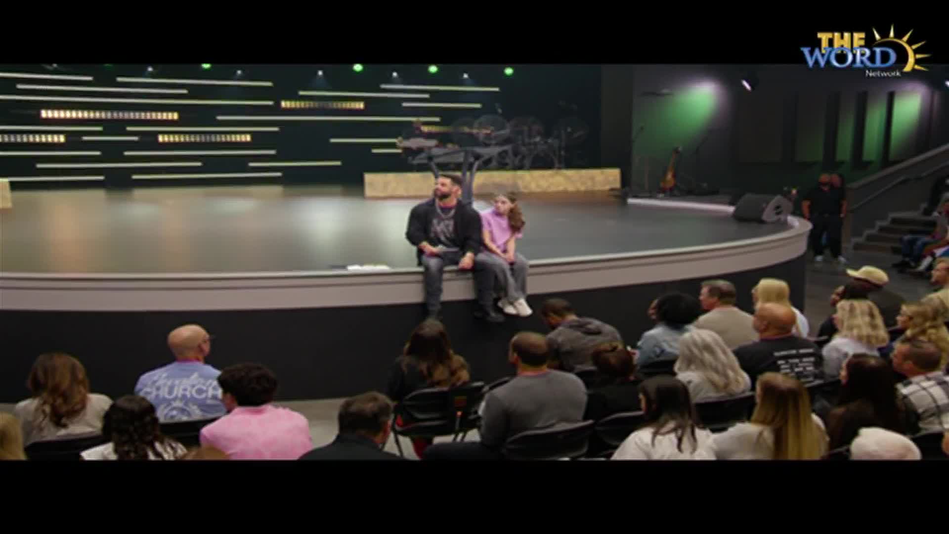 A man and a girl sit side-by-side on the edge of a stage, looking out at the audience. The Word Network logo is visible in the upper right corner.
