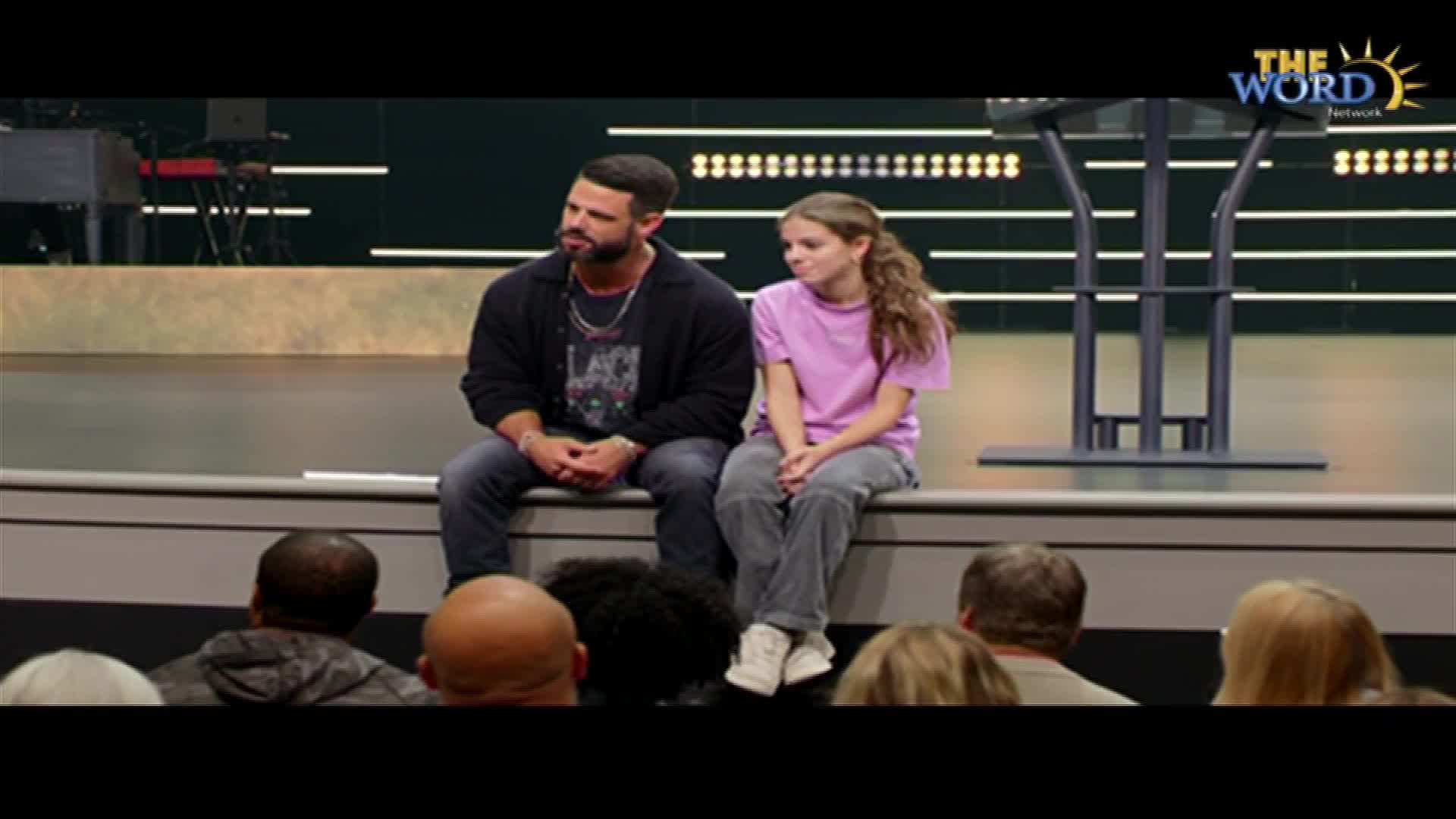 A man and a girl sit side-by-side on a stage, looking out at the audience. The Word Network logo is visible in the corner.
