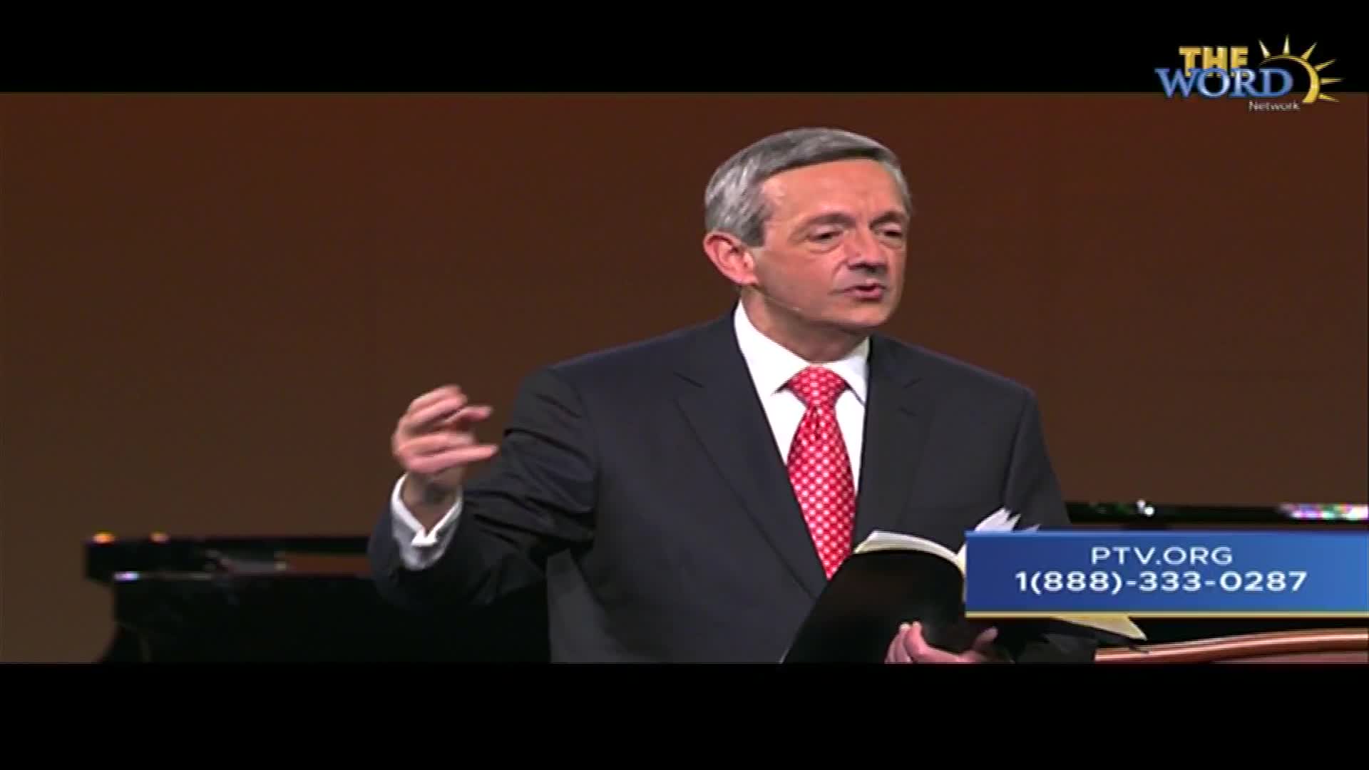 A man in a suit, speaking into a microphone, gestures with his left hand as he holds a book. The Word Network logo is visible in the upper right corner, and the PTV.org phone number is displayed at the bottom of the screen.
