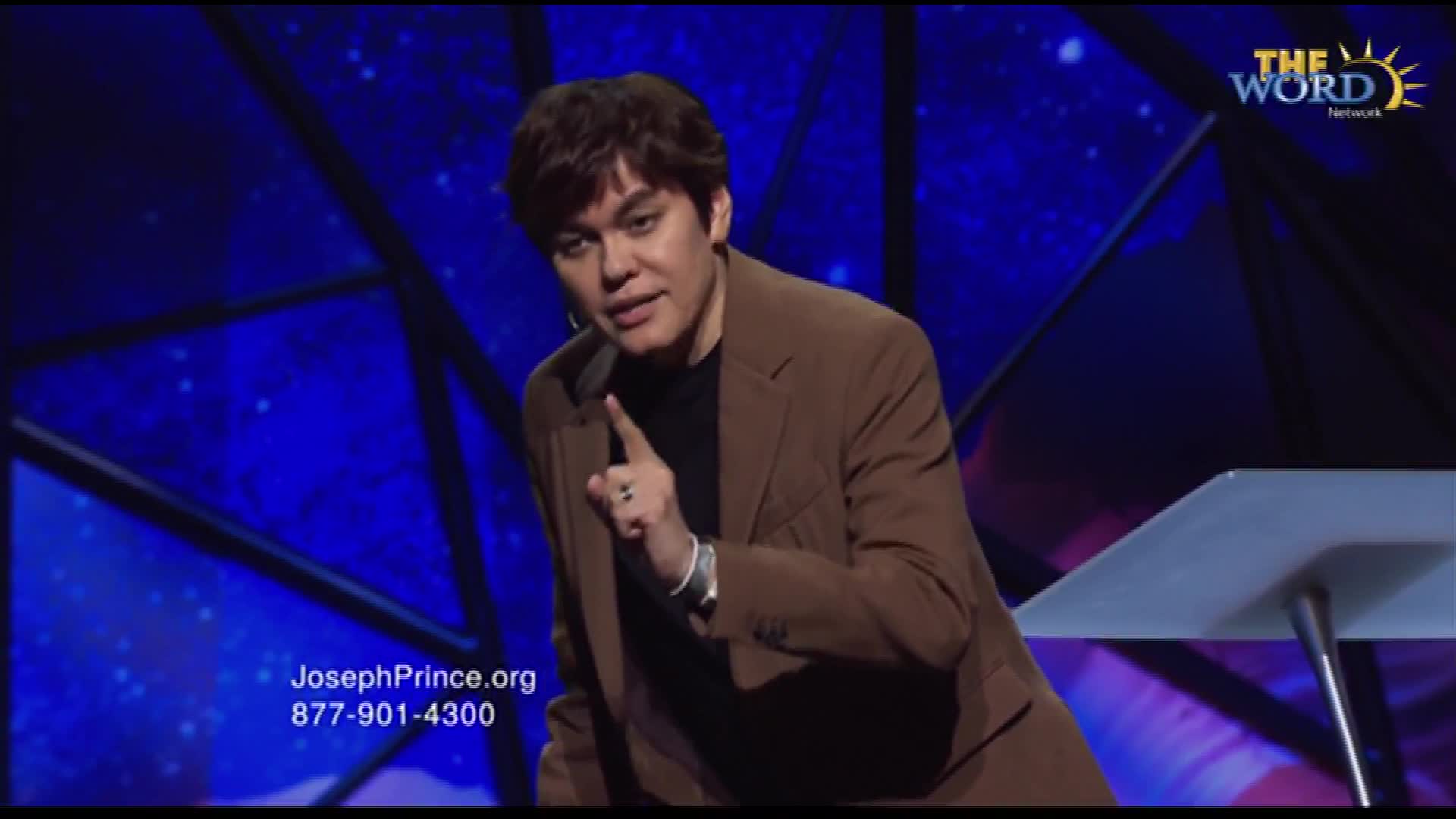 A man in a brown suit gestures emphatically, his index finger pointed directly at the camera. The backdrop is a deep blue, and the logo for The Word Network is visible in the upper right.
