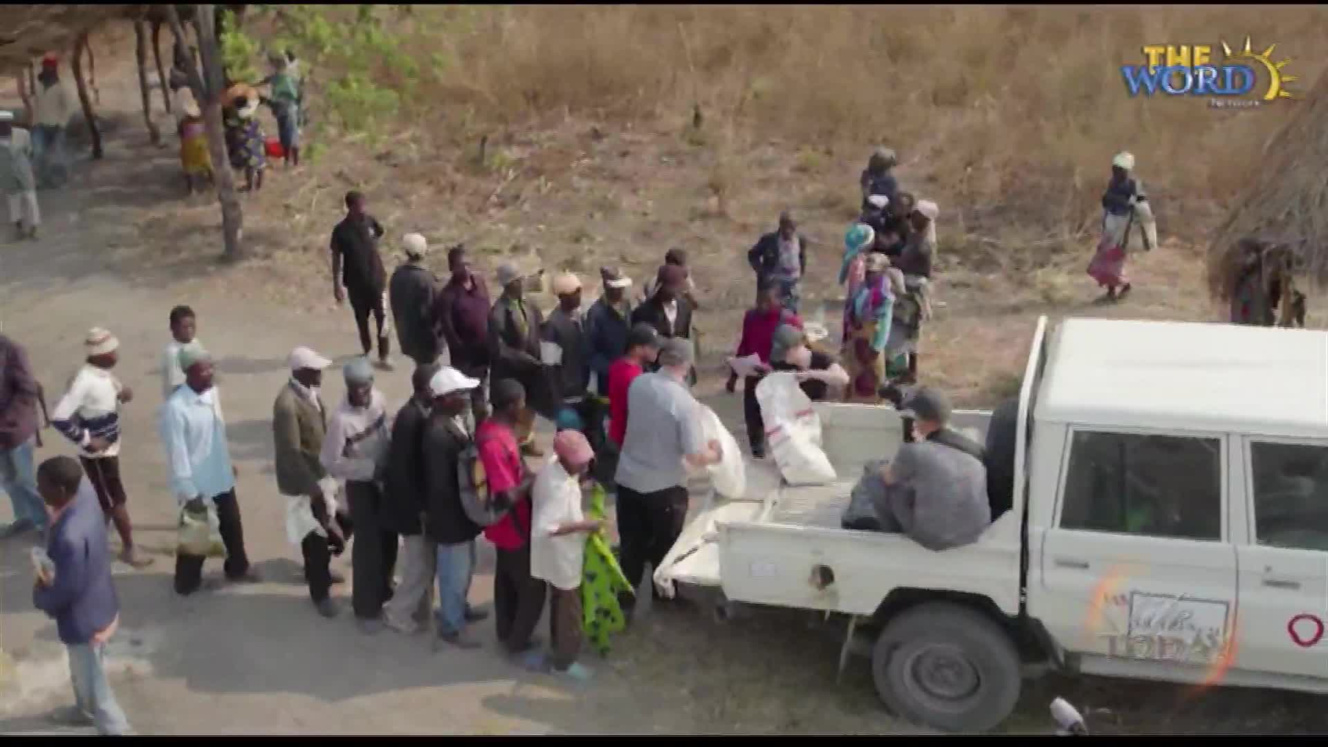 People wait patiently in a line, while others unload sacks from the back of a white truck. The Word Network logo is visible in the corner, suggesting this is a charitable effort.
