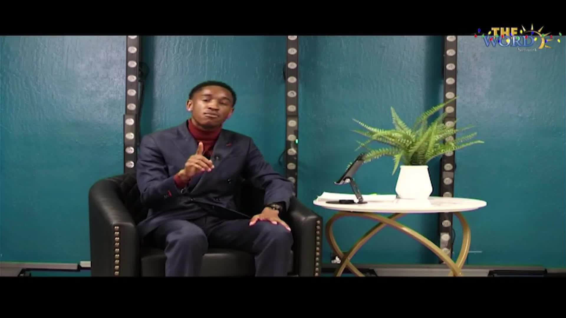 A man in a suit sits in a plush chair, gesturing with his right hand. Beside him, a small table holds a tablet and a potted fern, all set against a teal backdrop.

