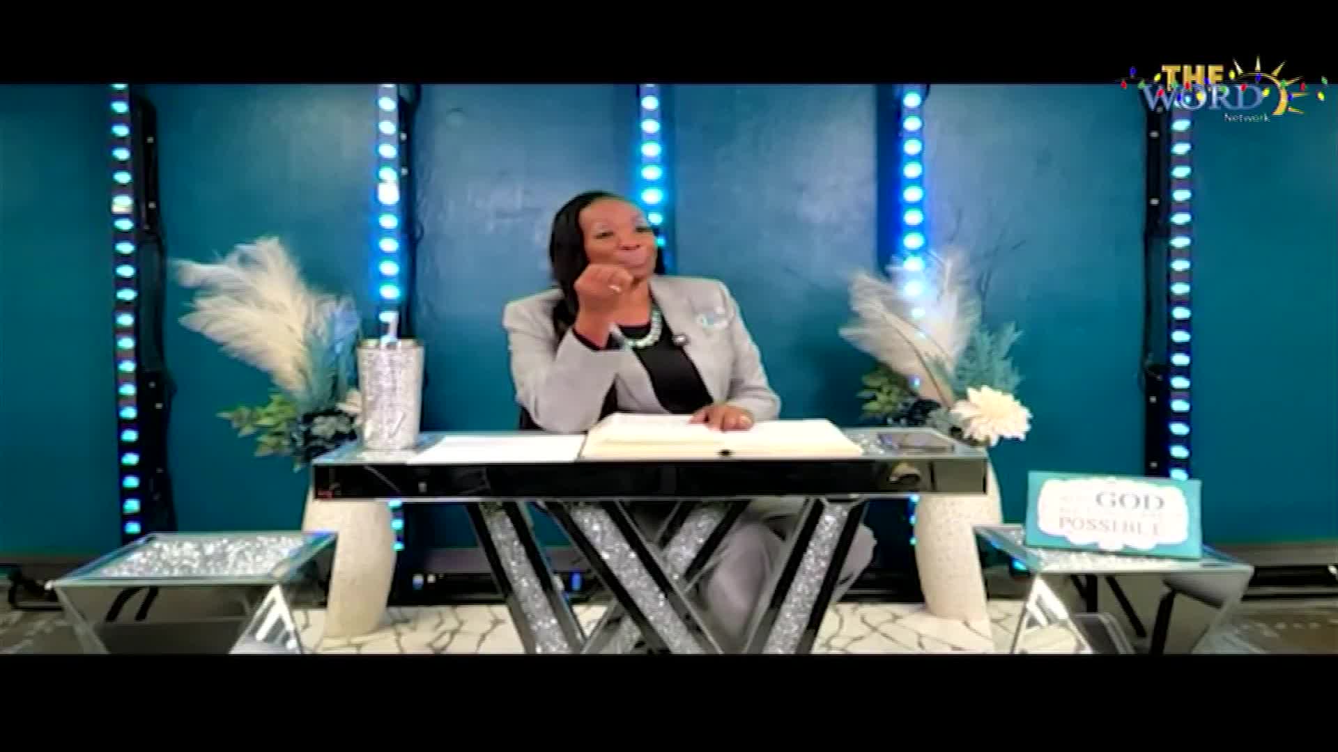 A woman in a gray suit sits at a mirrored desk, looking off to the side. Behind her, the logo for The Word Network is visible.
