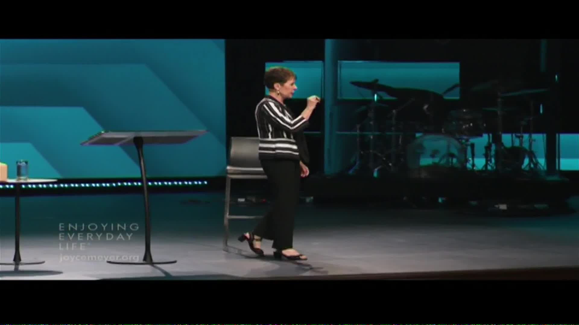 A woman in a striped shirt and black pants walks across a stage, gesturing with her right hand. Behind her, a drum set sits in the shadows.
